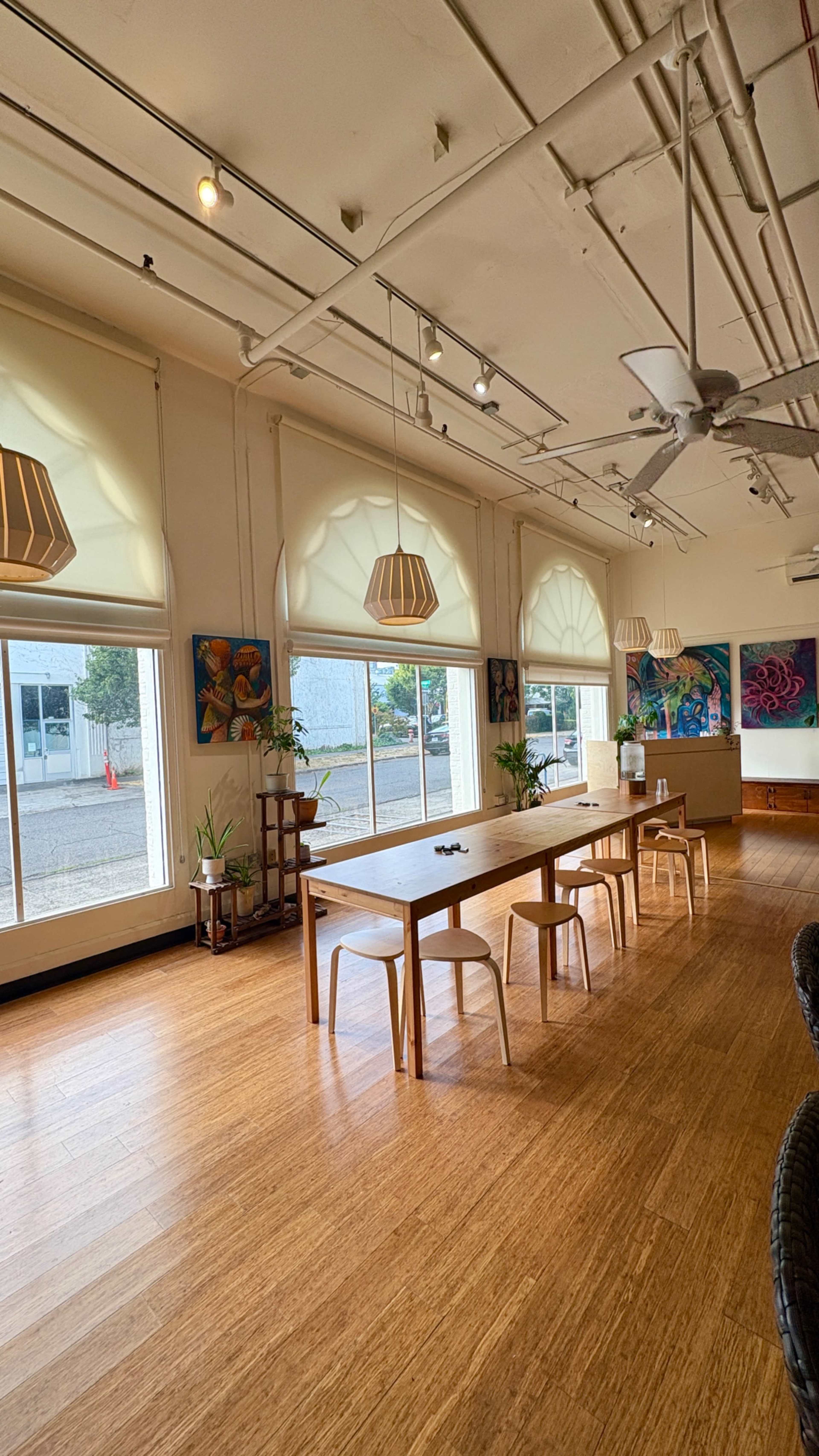 The image shows a spacious, well-lit room with large windows, featuring a long wooden table and several plants, adorned with artwork on the walls.