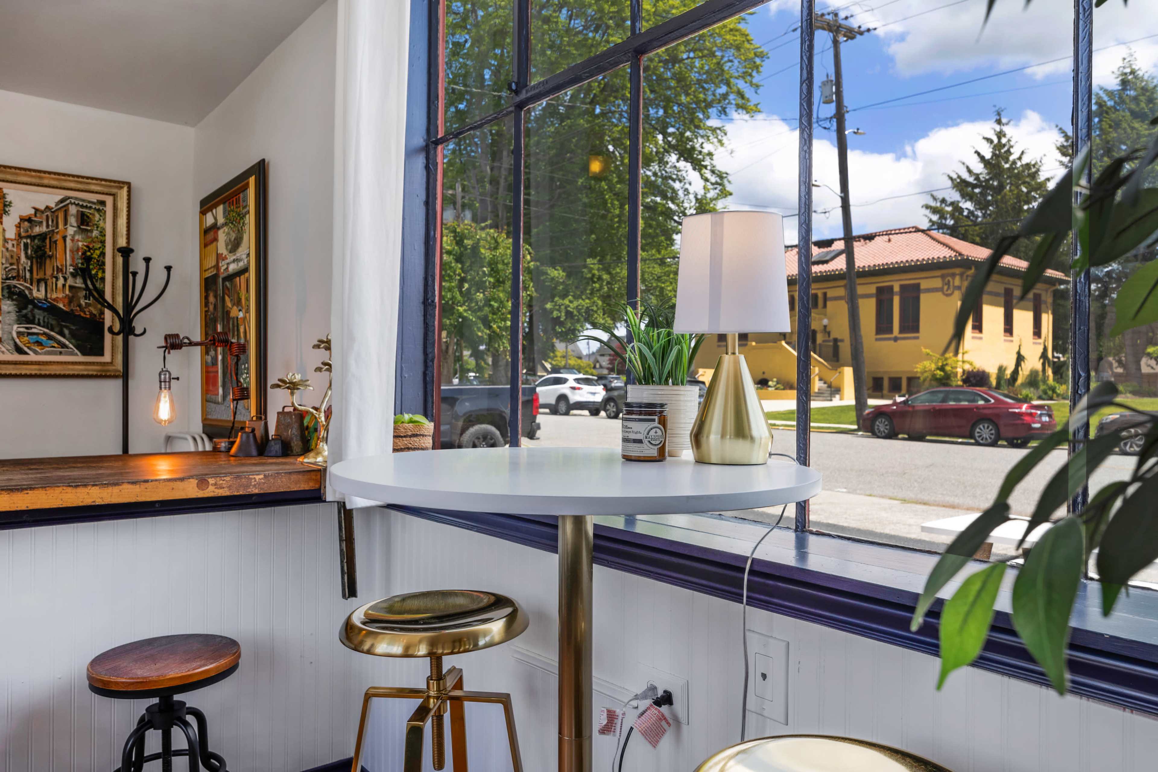 A bright, modern interior features a small round table with gold accents, surrounded by wooden stools, and a large window that opens to a view of a street with parked cars and a yellow building.