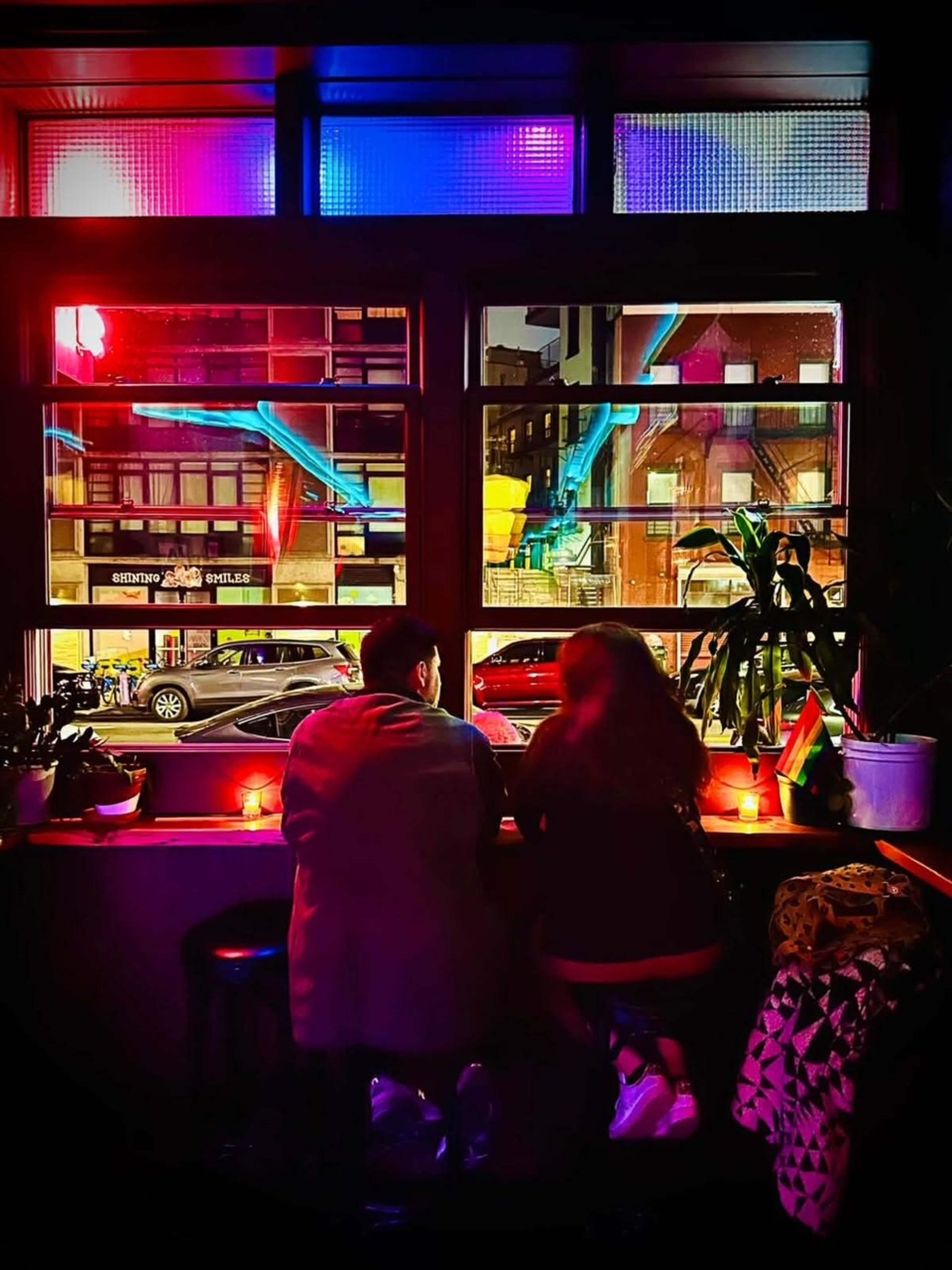 A couple sits at a window bar in a dimly lit venue, overlooking a colorful street scene illuminated by neon lights.