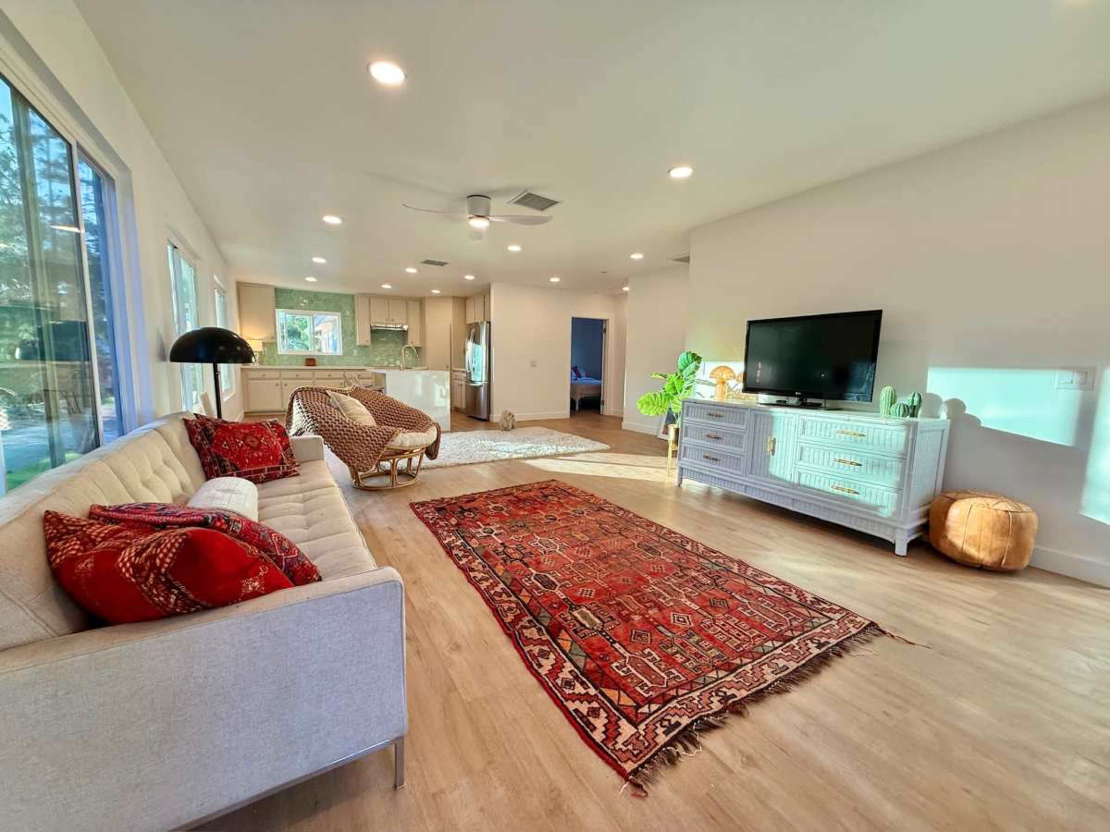 A modern living room features a light-colored sofa, a patterned area rug, a television on a white console, and large windows allowing natural light to fill the space.