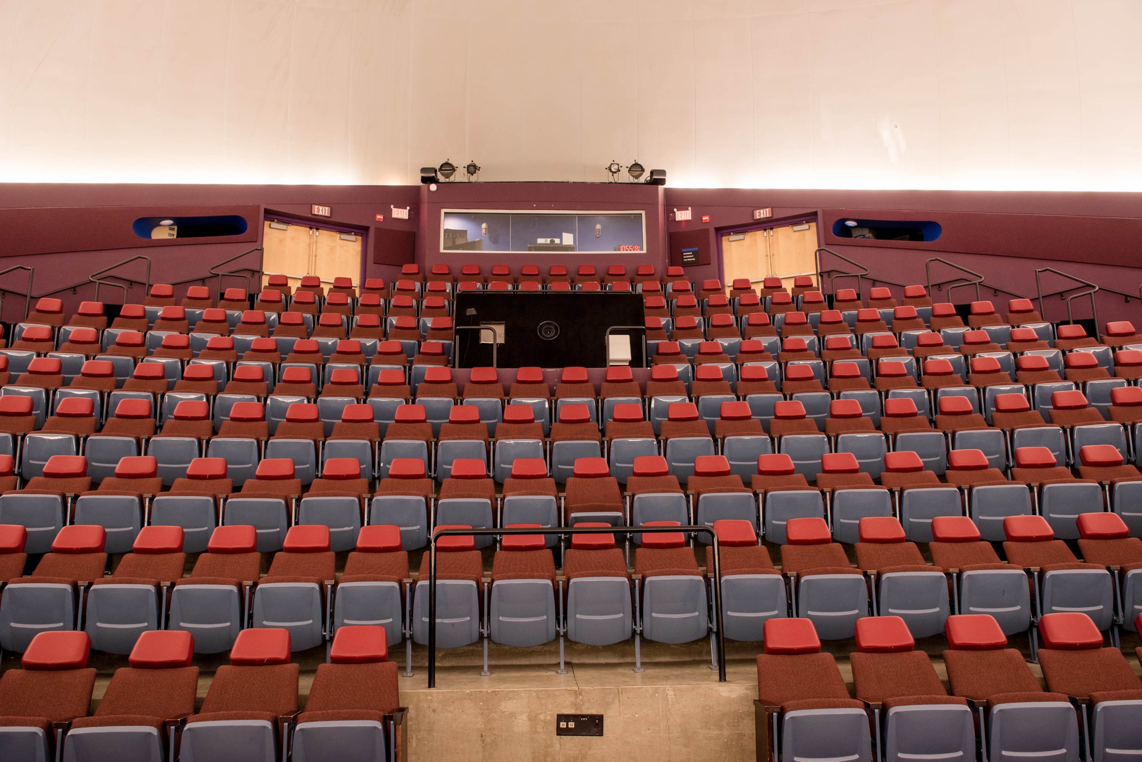 The image shows an empty theater with rows of red and blue seats arranged in a semicircular layout facing a stage.