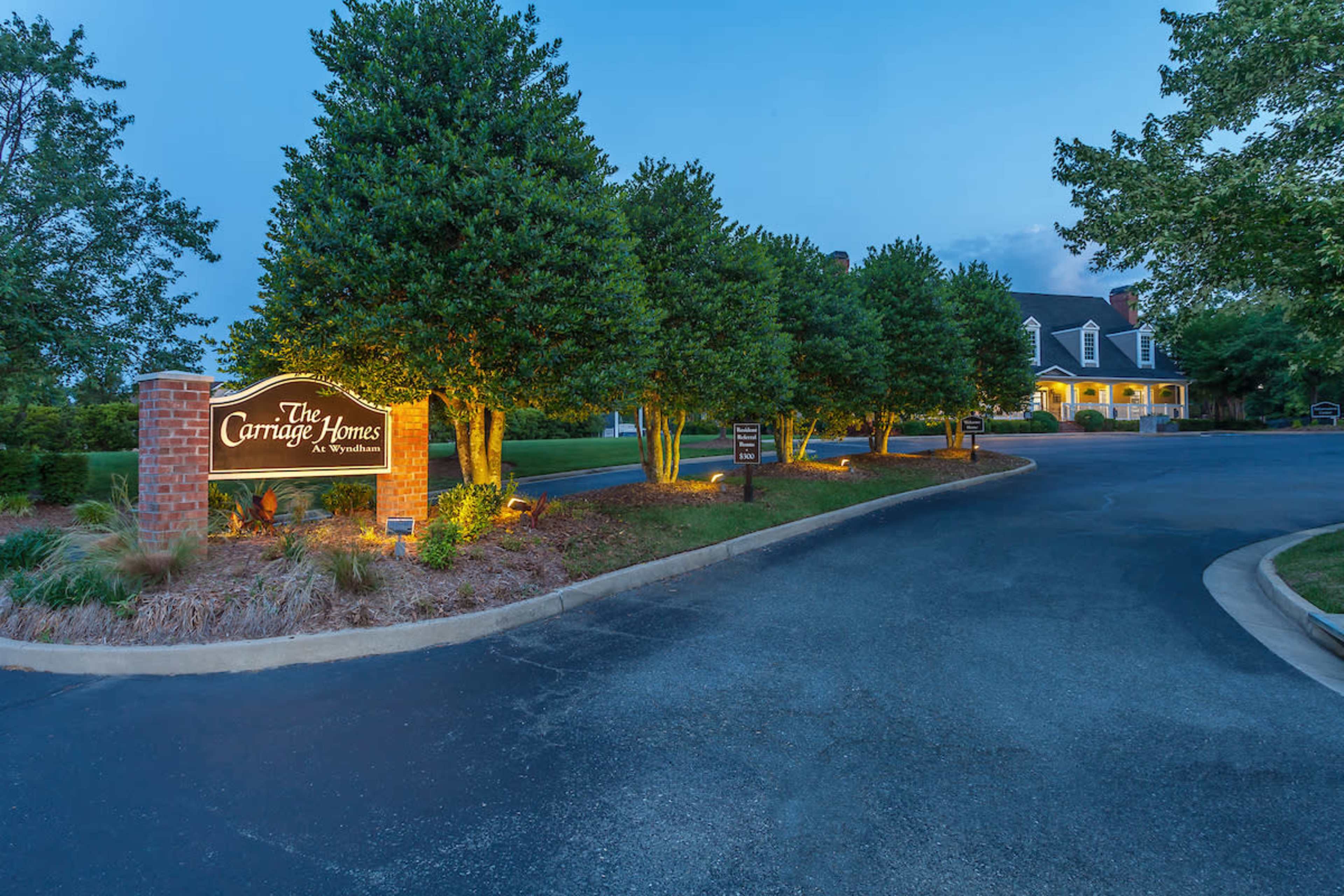 A well-lit entrance to The Carriage Homes with neatly trimmed trees along a paved road leading to a building in the background.