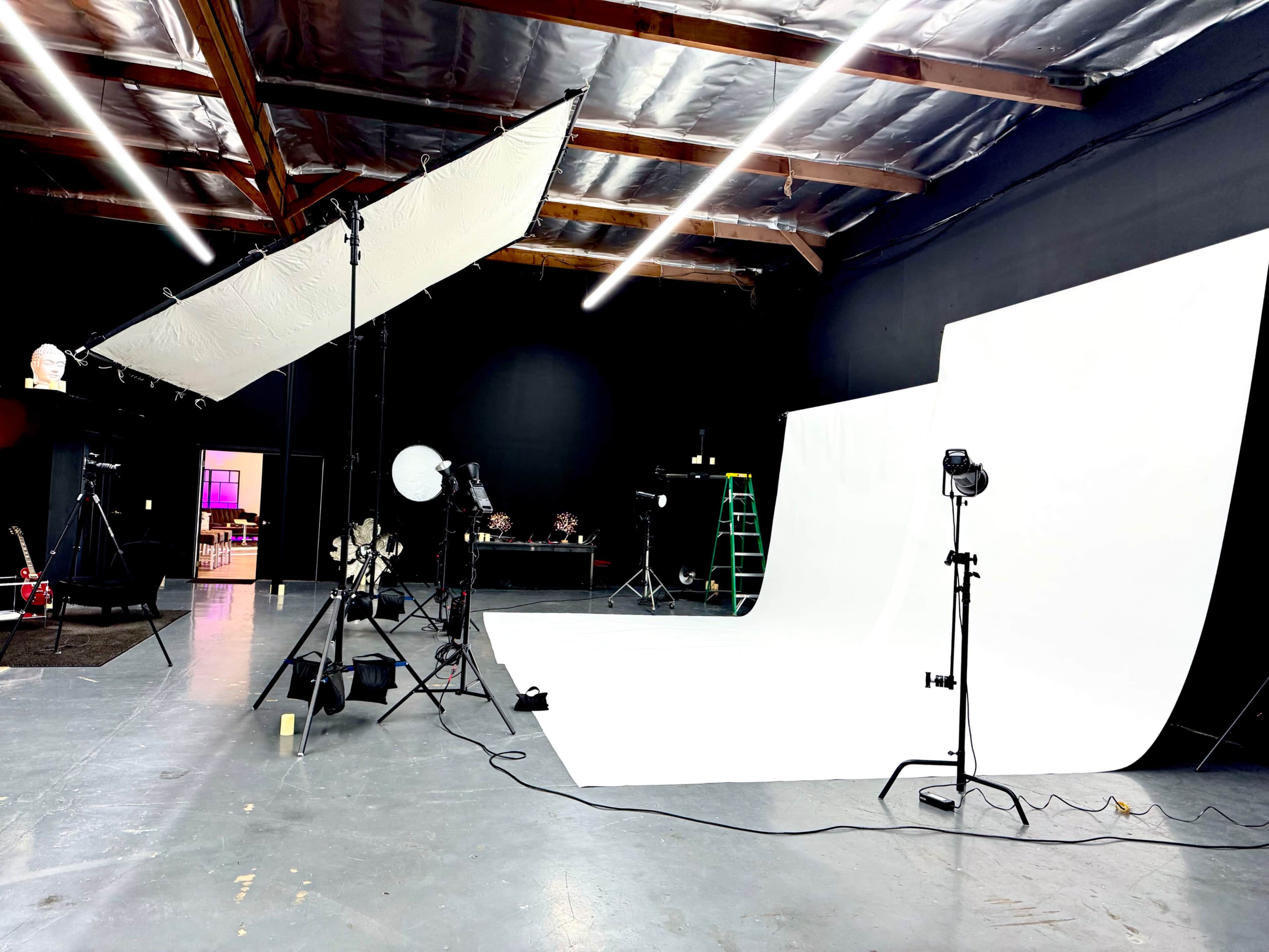 The image shows a photography studio with a white backdrop, various lighting equipment, and a ladder positioned against the wall.