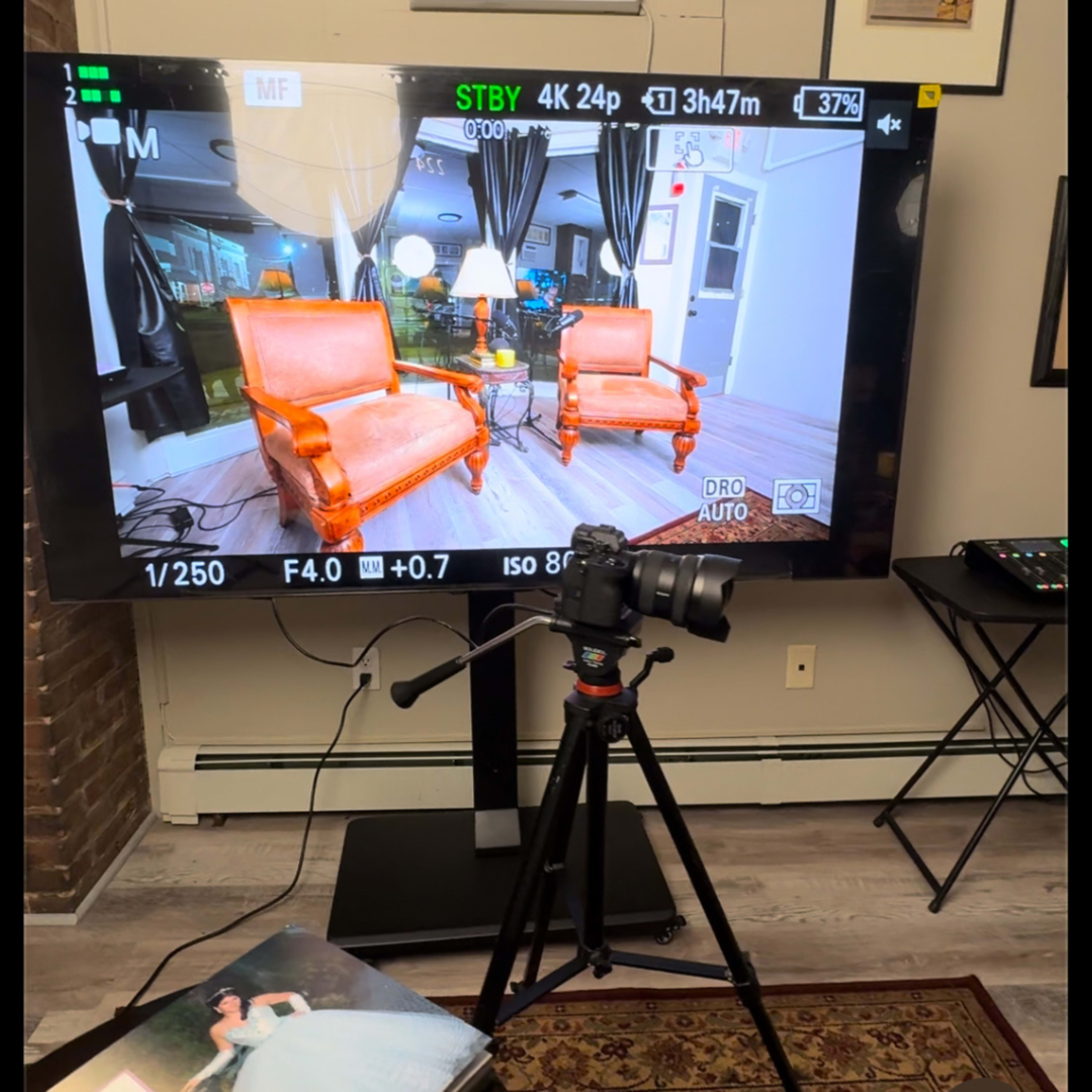 A camera on a tripod captures the image of a room featuring two orange chairs and various decorative elements, displayed on a large monitor.