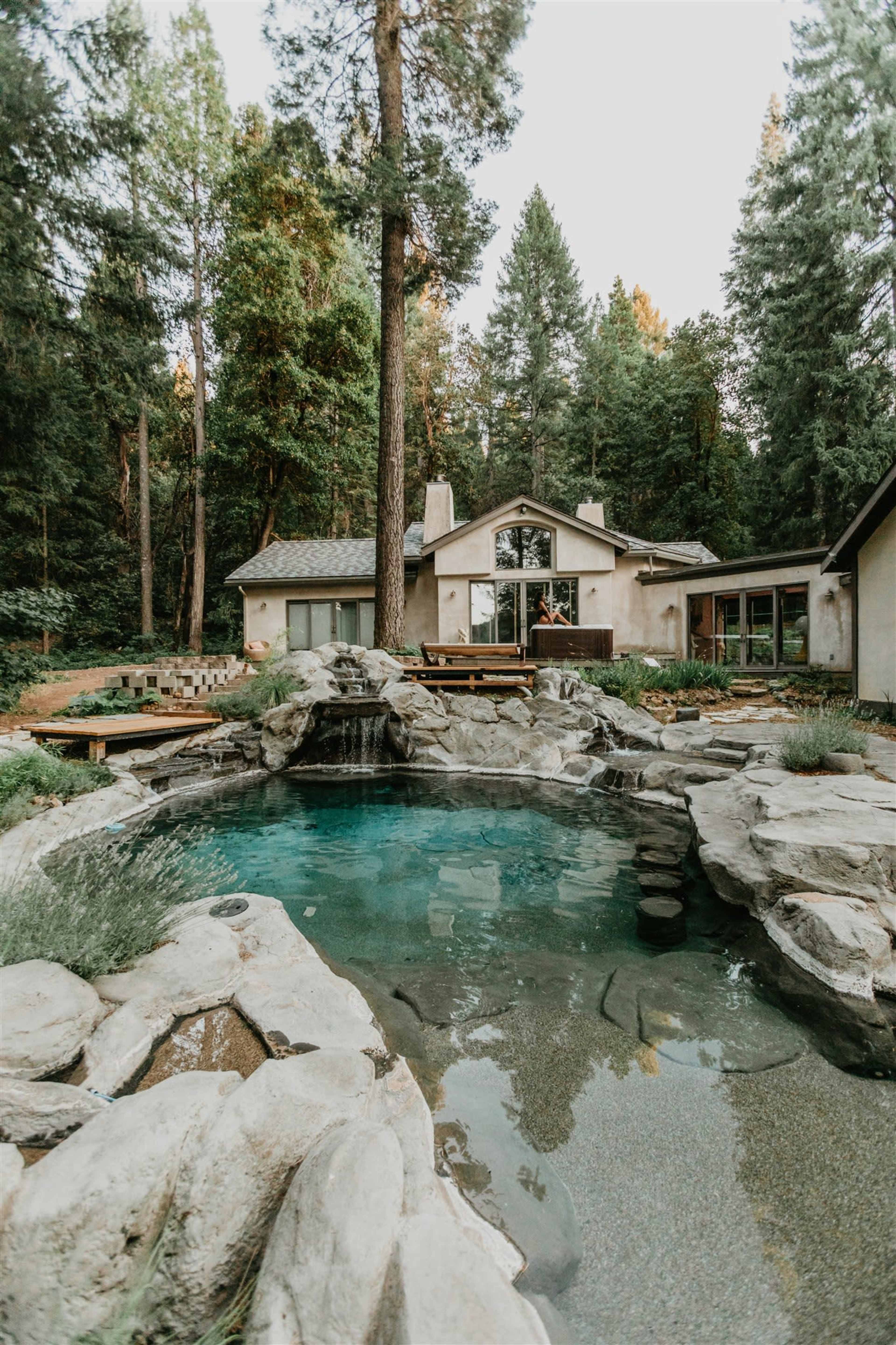 A rocky pool is surrounded by natural landscaping and a cabin-like house set among tall pine trees.