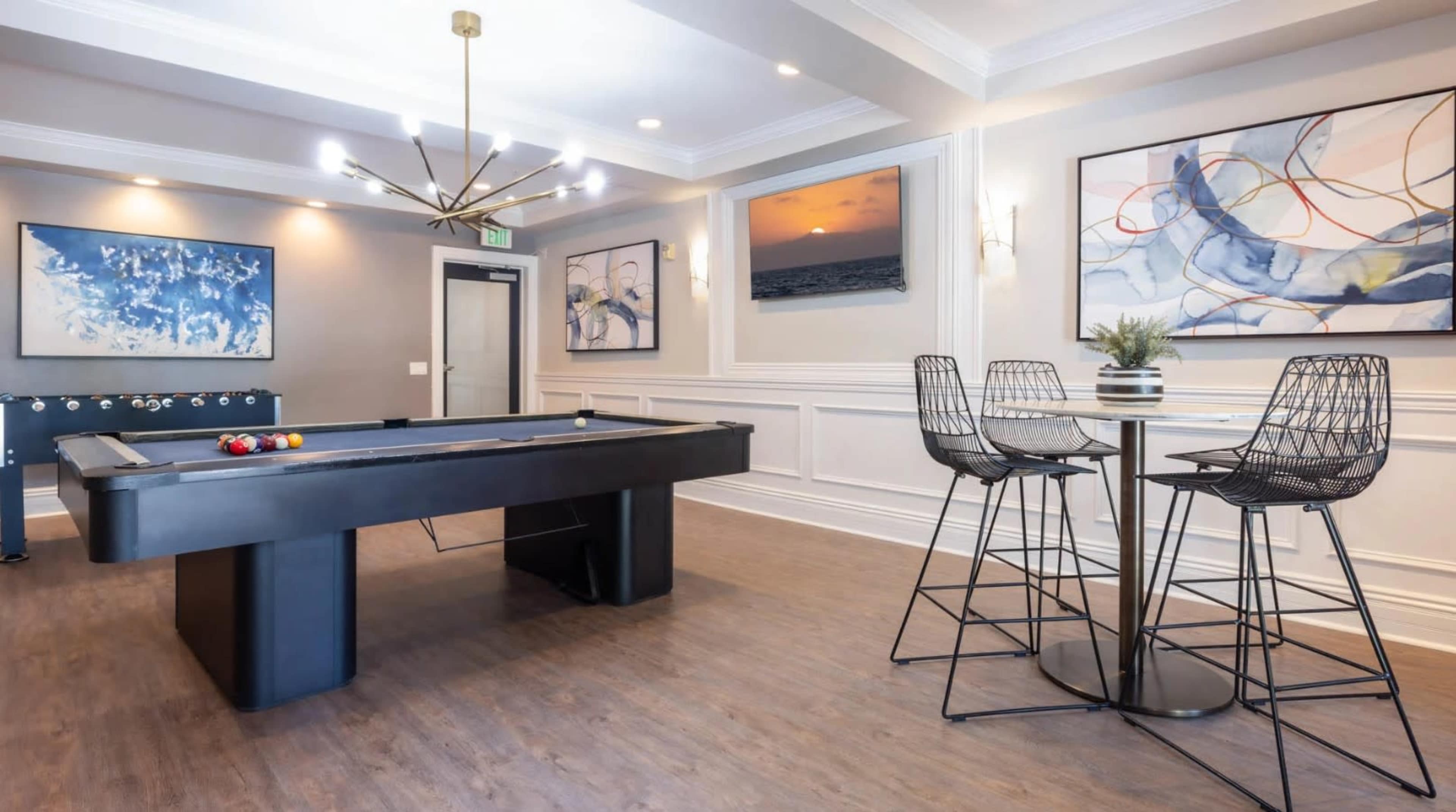 The image shows a modern game room featuring a pool table, bar-height seating, and framed artwork on the walls.