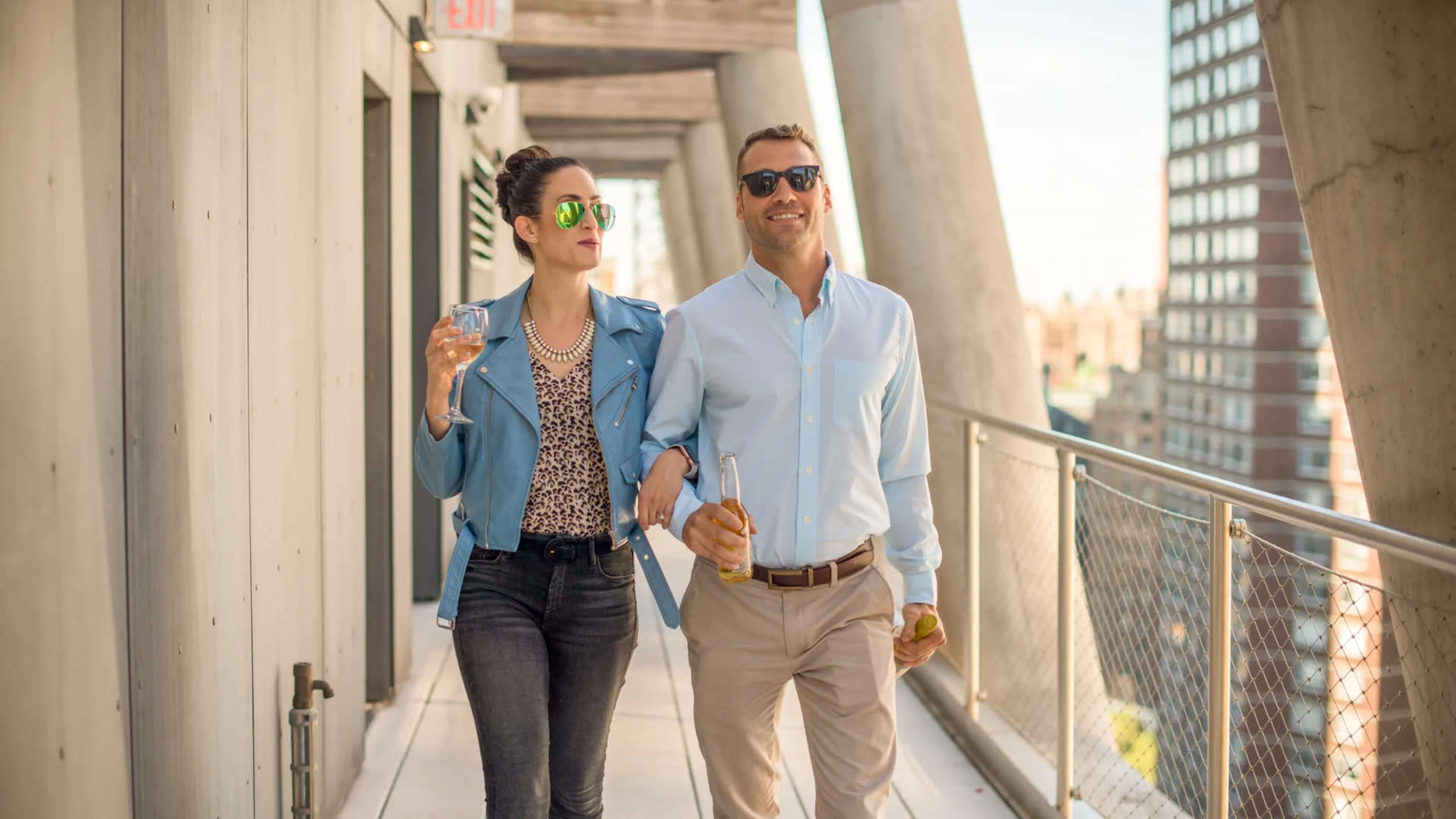 A man and a woman walk arm in arm on a balcony, both holding drinks and wearing sunglasses.