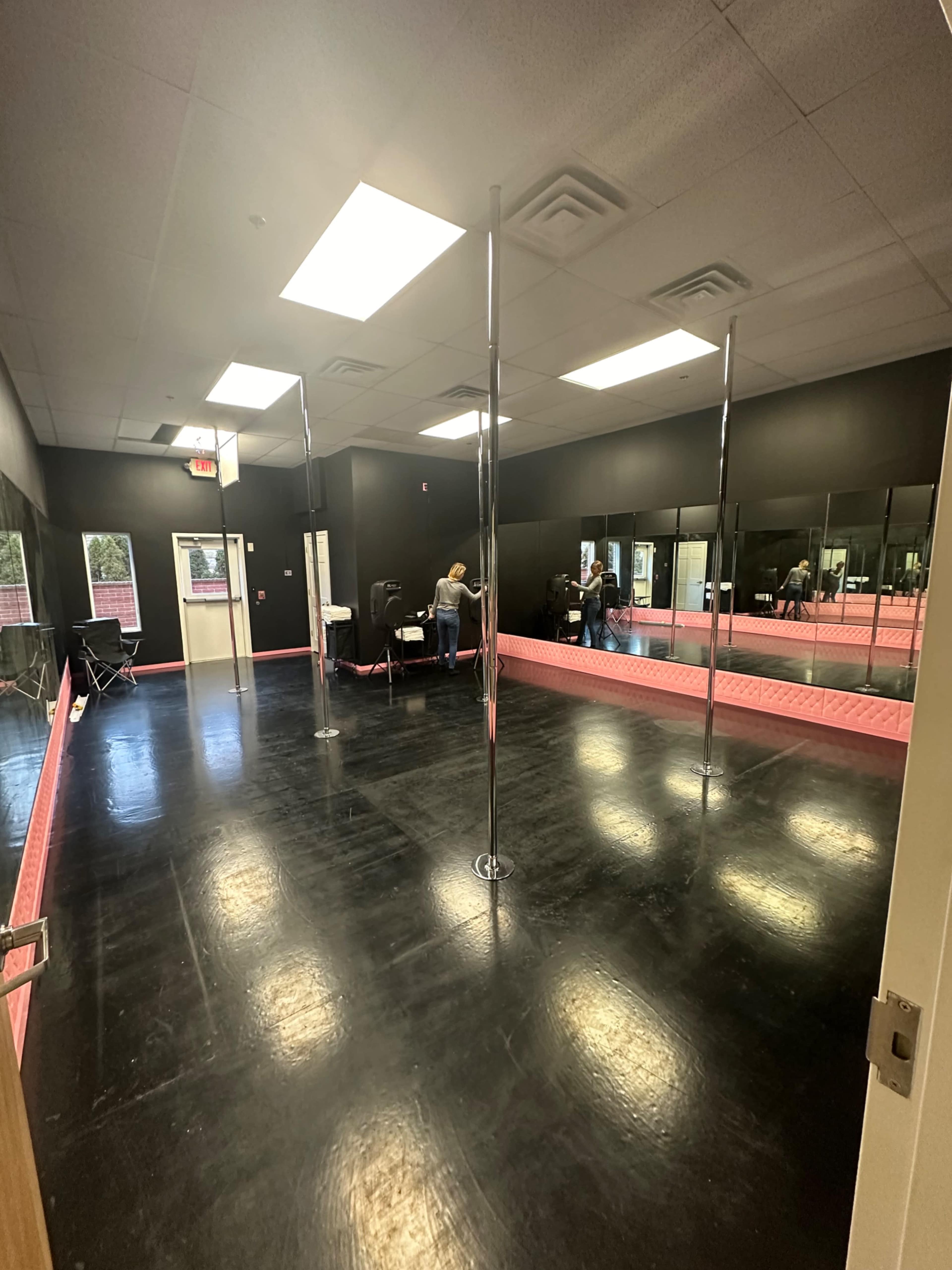 The image shows a dance studio with black walls, a polished floor, and several poles arranged in an open space, featuring mirrors along one side.