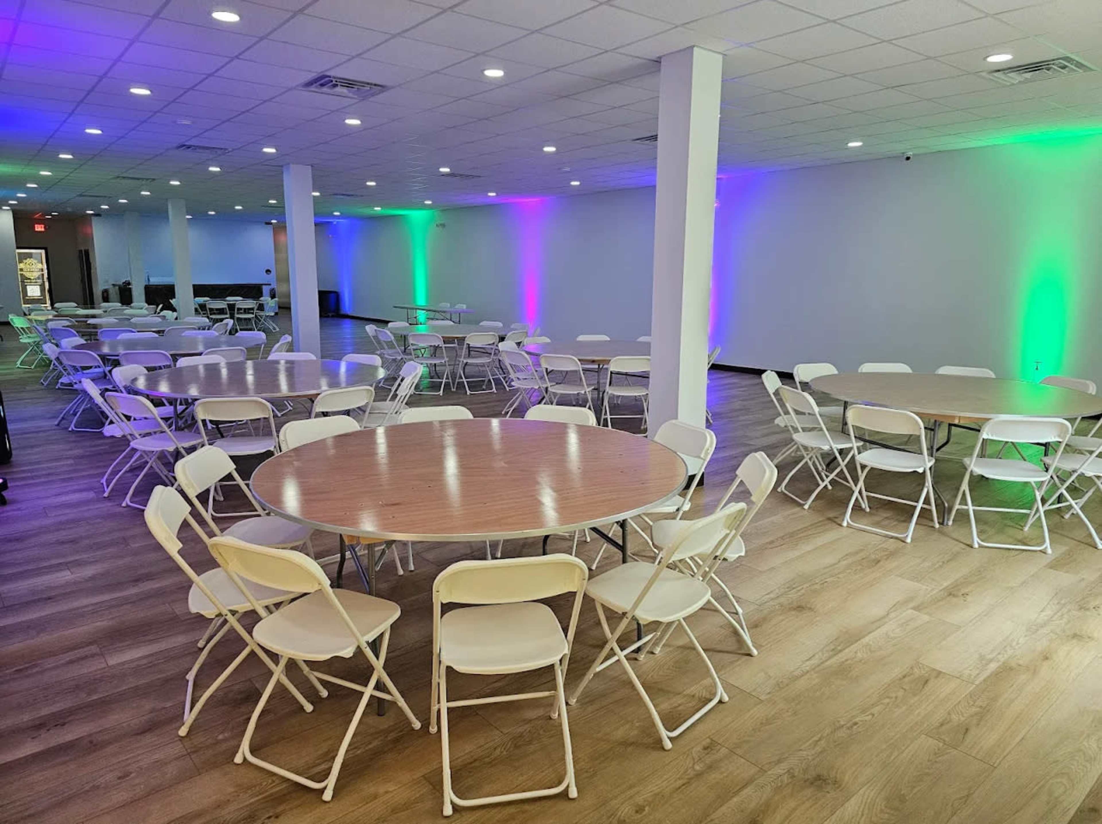 The image shows a spacious, well-lit event room with round tables and white folding chairs arranged neatly on a wooden floor, illuminated by colored lights.