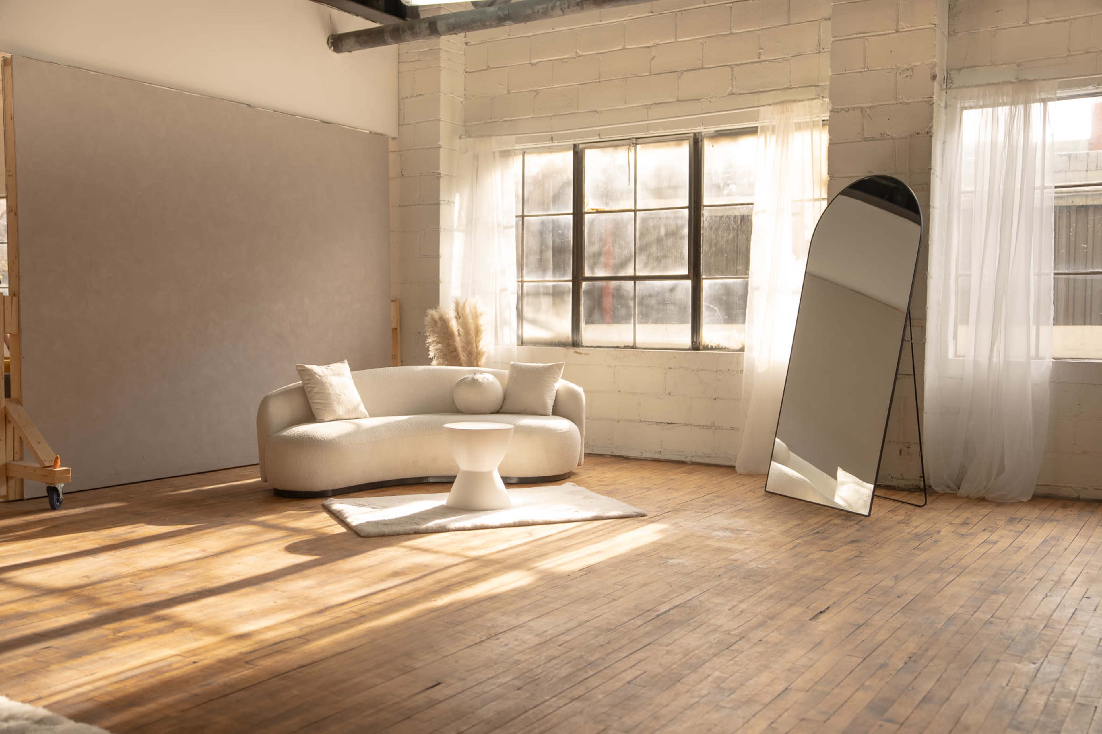 The image shows a bright, modern interior featuring a curved sofa, a small table, a large mirror, and a window with natural light streaming in.