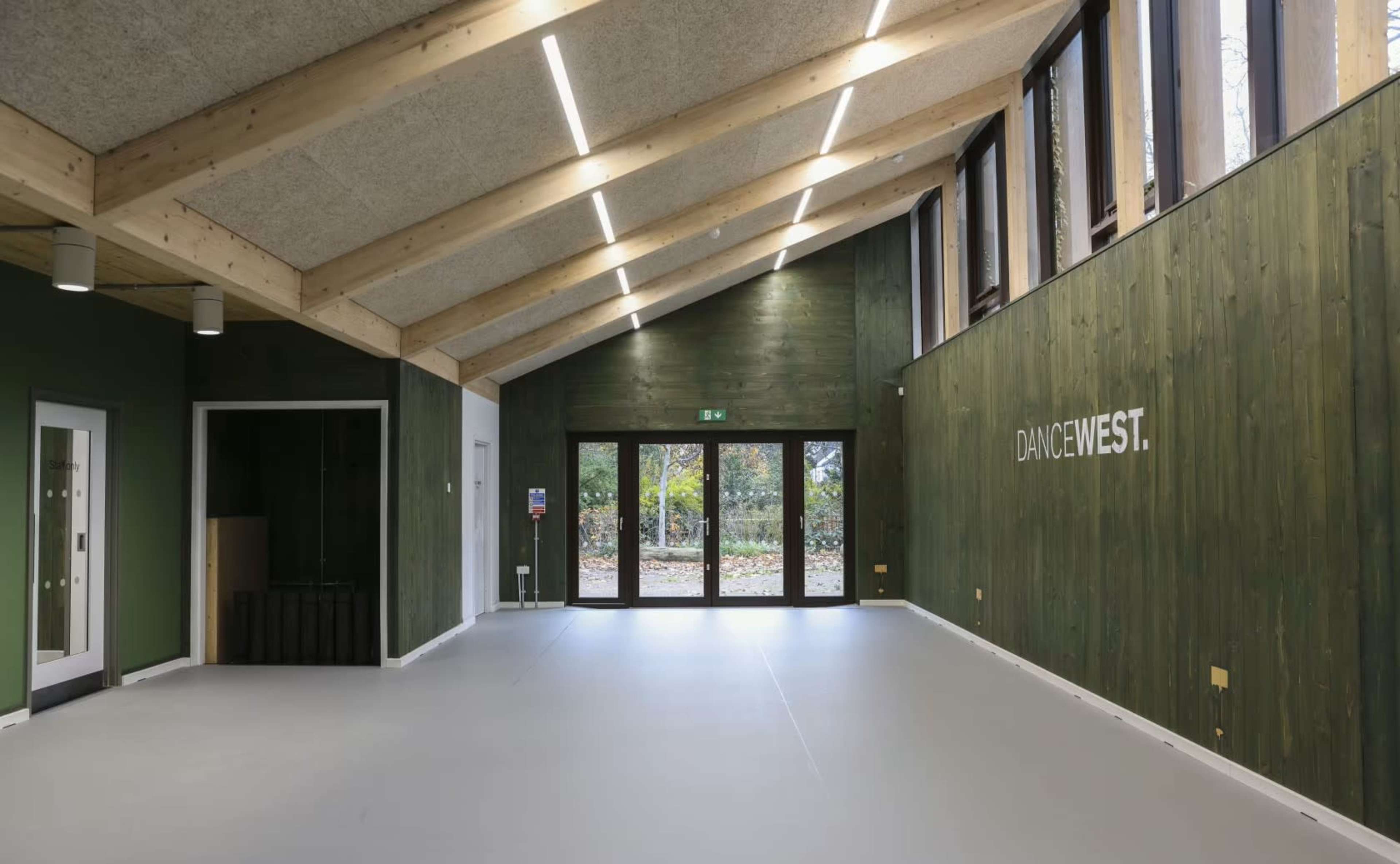 The image depicts an empty dance studio featuring high wooden ceilings, green walls, and large windows looking out onto a garden area.
