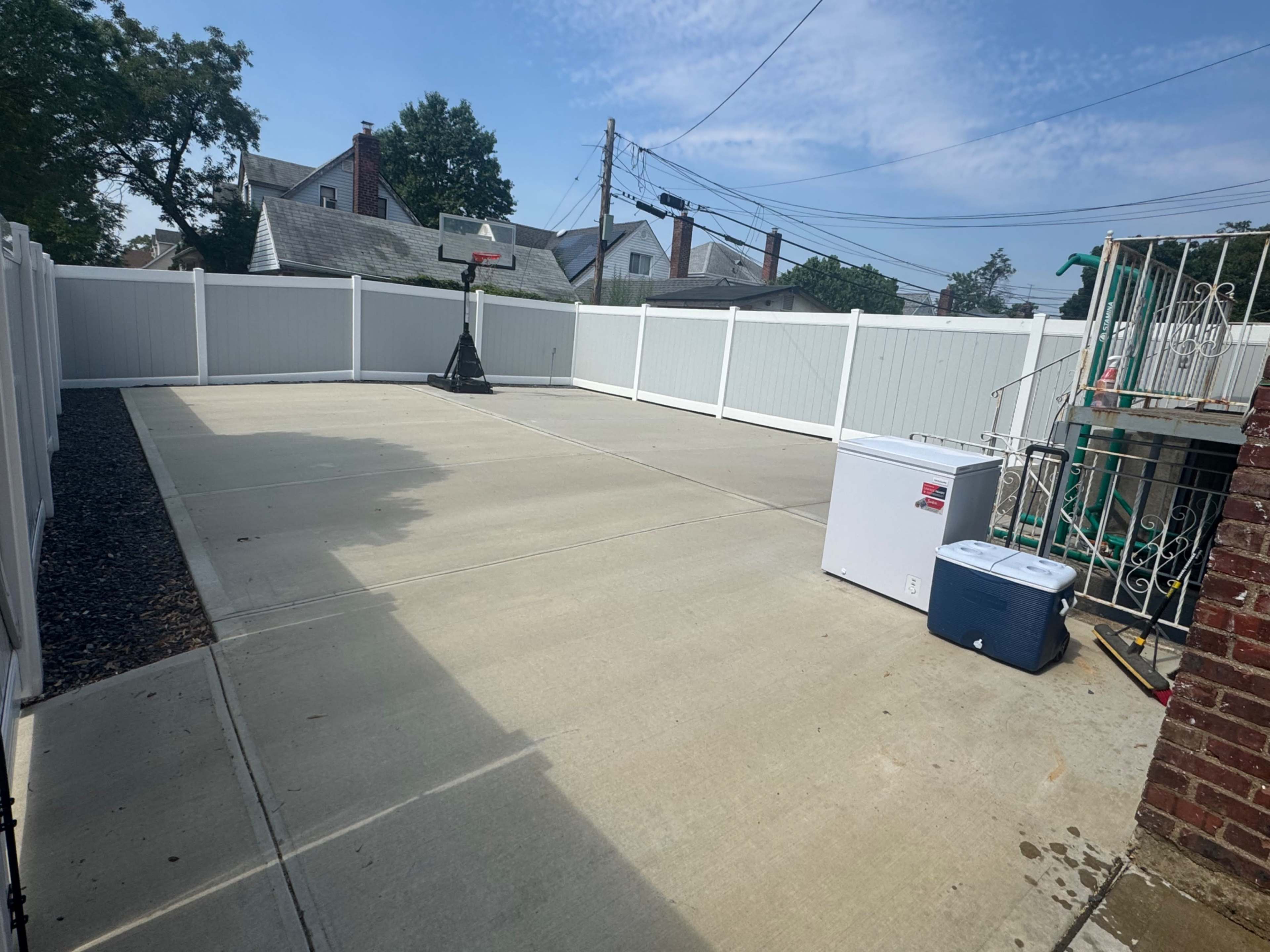 The image shows a paved outdoor area with a basketball hoop, a cooler, and a small white appliance, enclosed by a white fence.