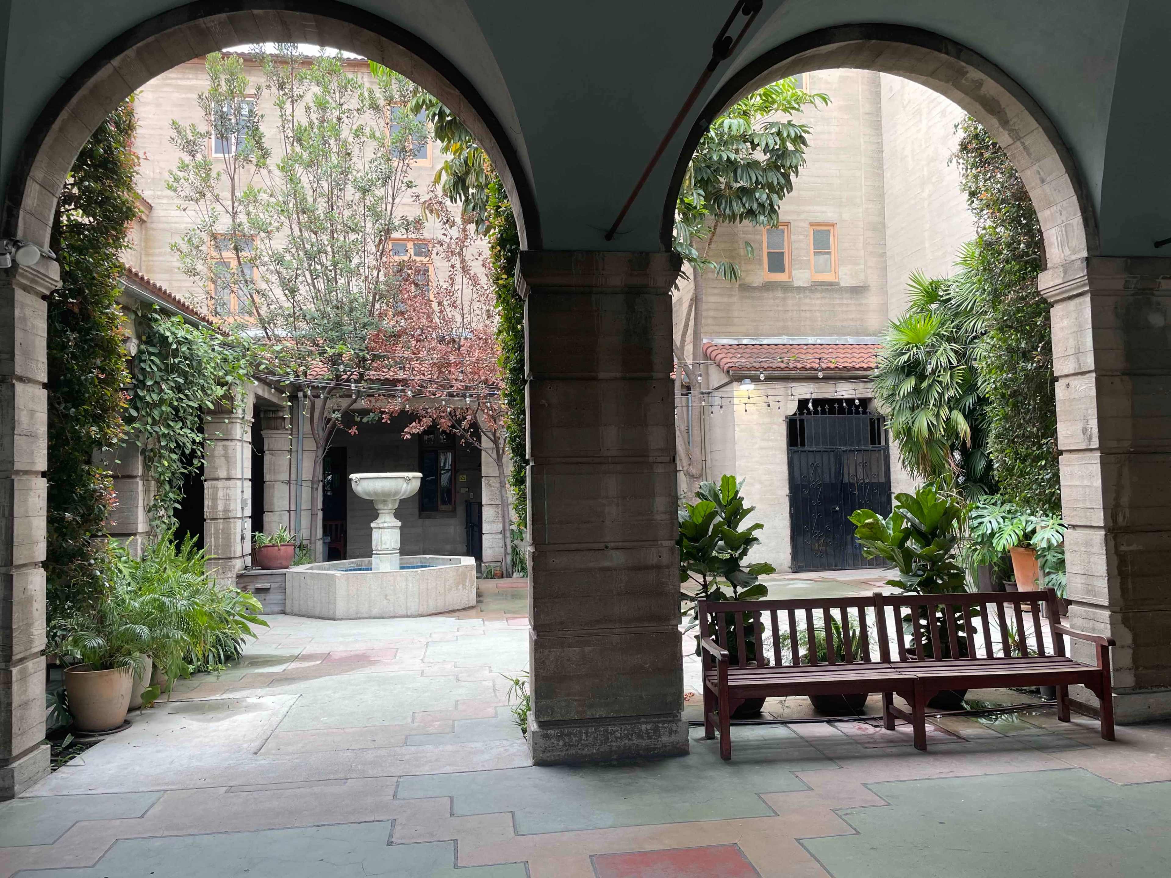 The image shows a courtyard with arches, a stone fountain, potted plants, and a wooden bench amidst greenery and a building facade.