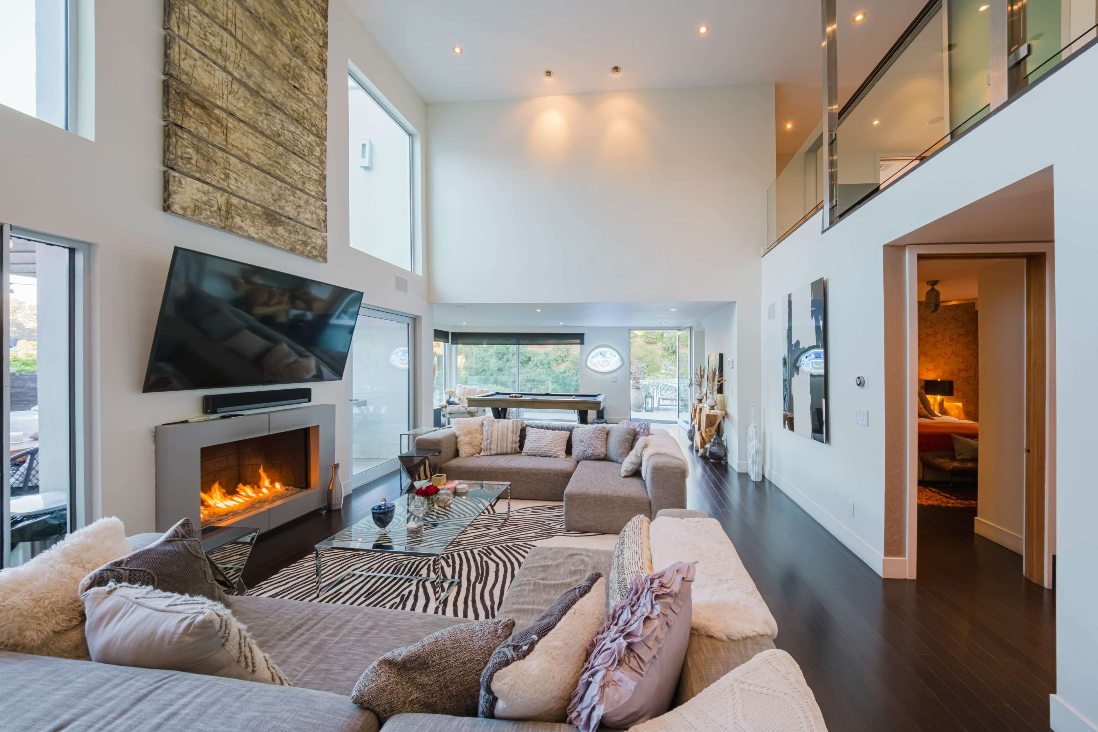The image shows a spacious, modern living room featuring a large sectional sofa, a central fireplace, and floor-to-ceiling windows that allow natural light to fill the space.