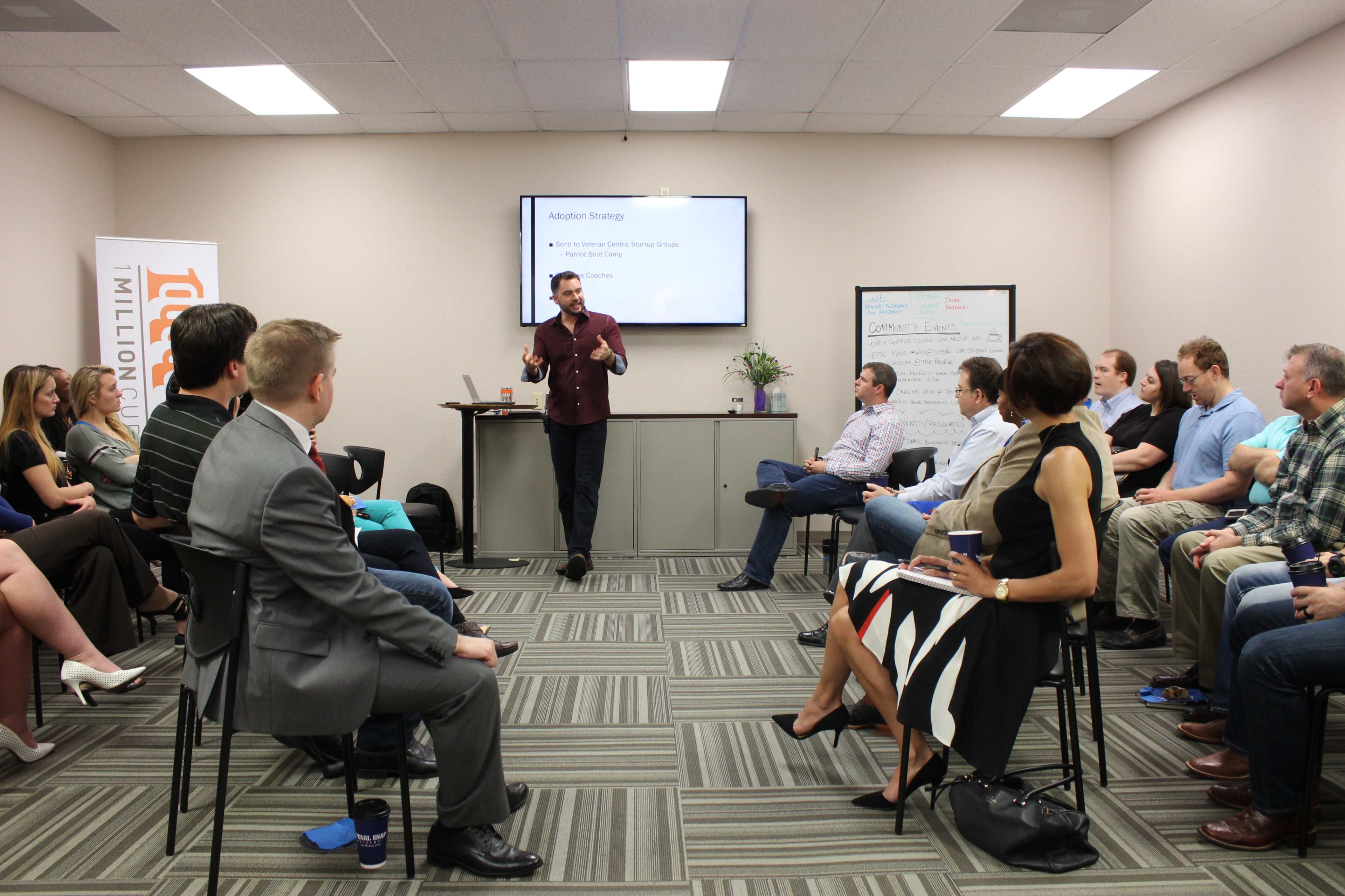 A speaker presents to an audience seated in a meeting room with a projector screen displaying information.