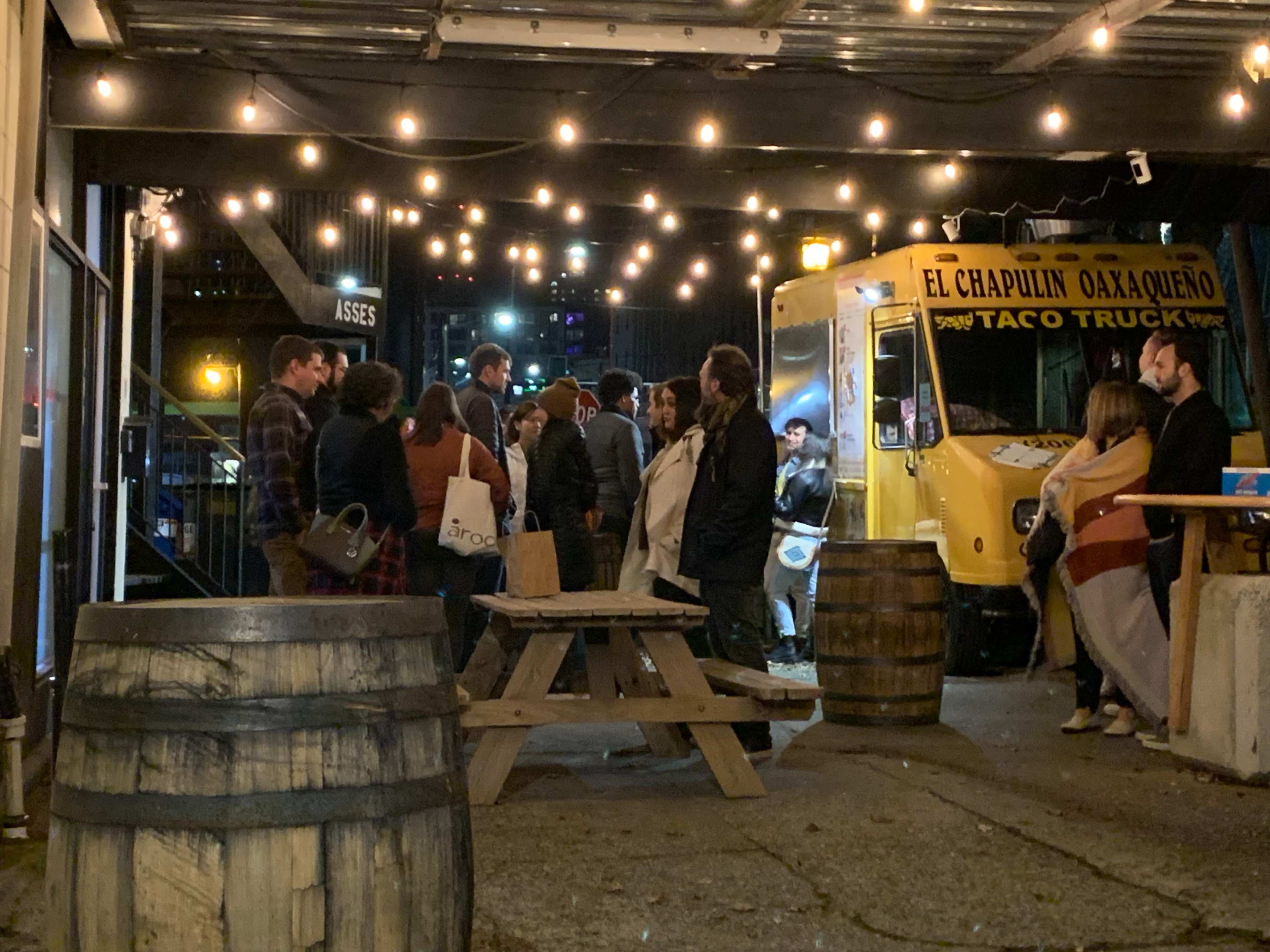 A group of people gathers around a yellow taco truck under string lights in a courtyard with wooden tables and barrels.
