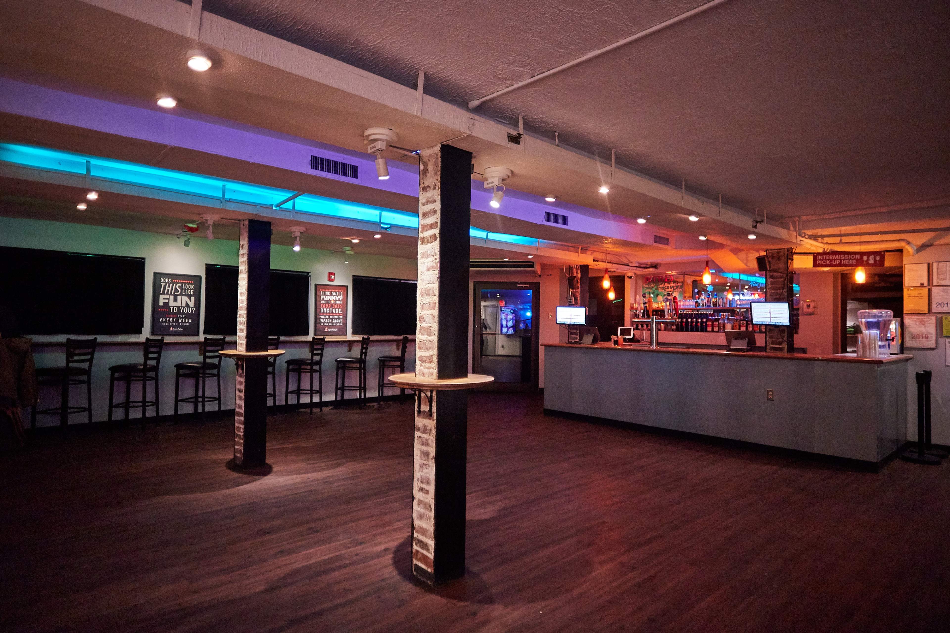 An empty bar interior features a counter with colorful lighting, exposed brick pillars, and seating arranged against the walls.