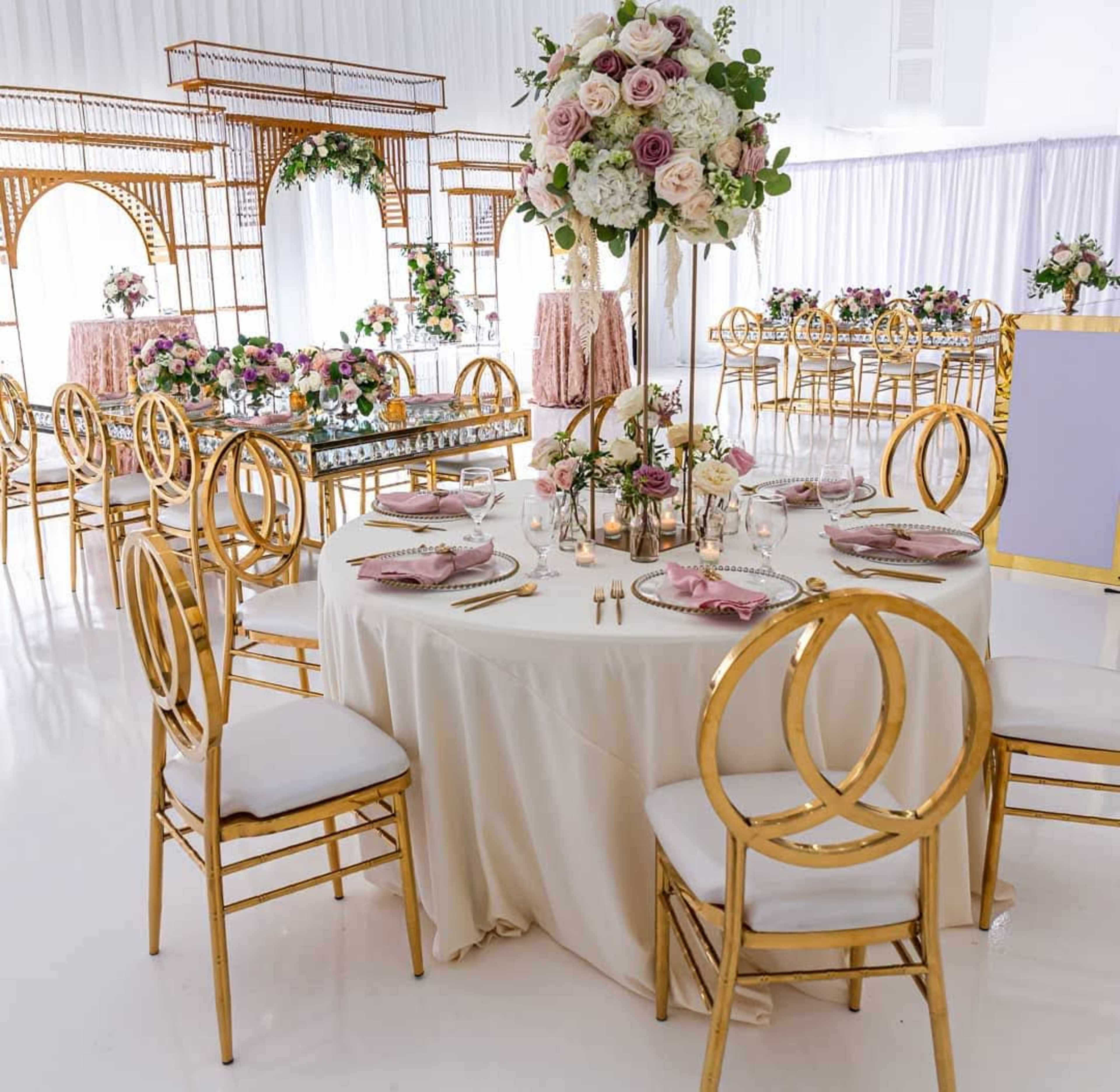 A dining setup features elegantly arranged tables with gold chairs, floral centerpieces, and pastel-colored tableware in a bright venue.