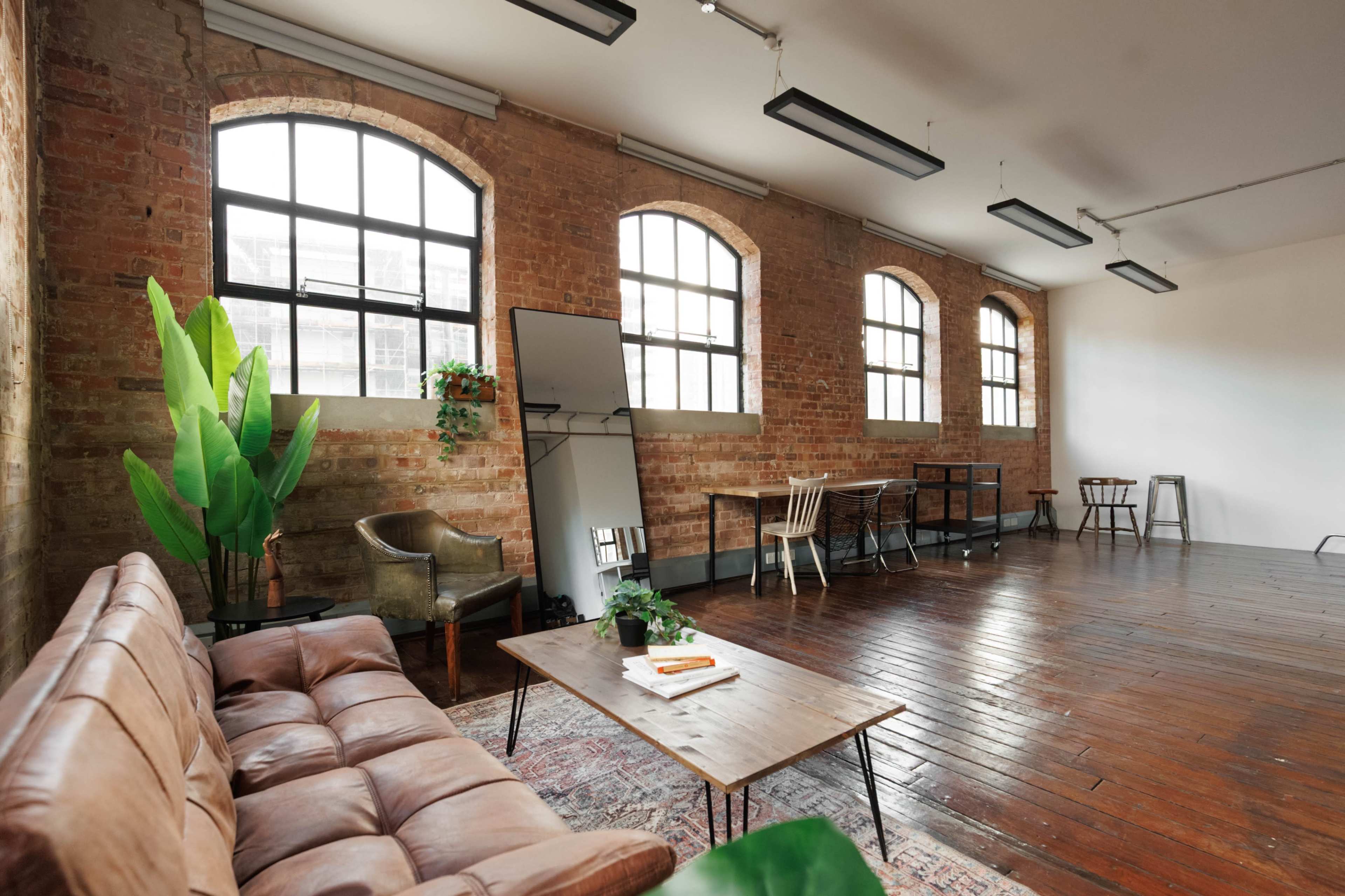 The image shows a spacious, industrial-style room with exposed brick walls, large windows, a leather sofa, a wooden table, and plants.