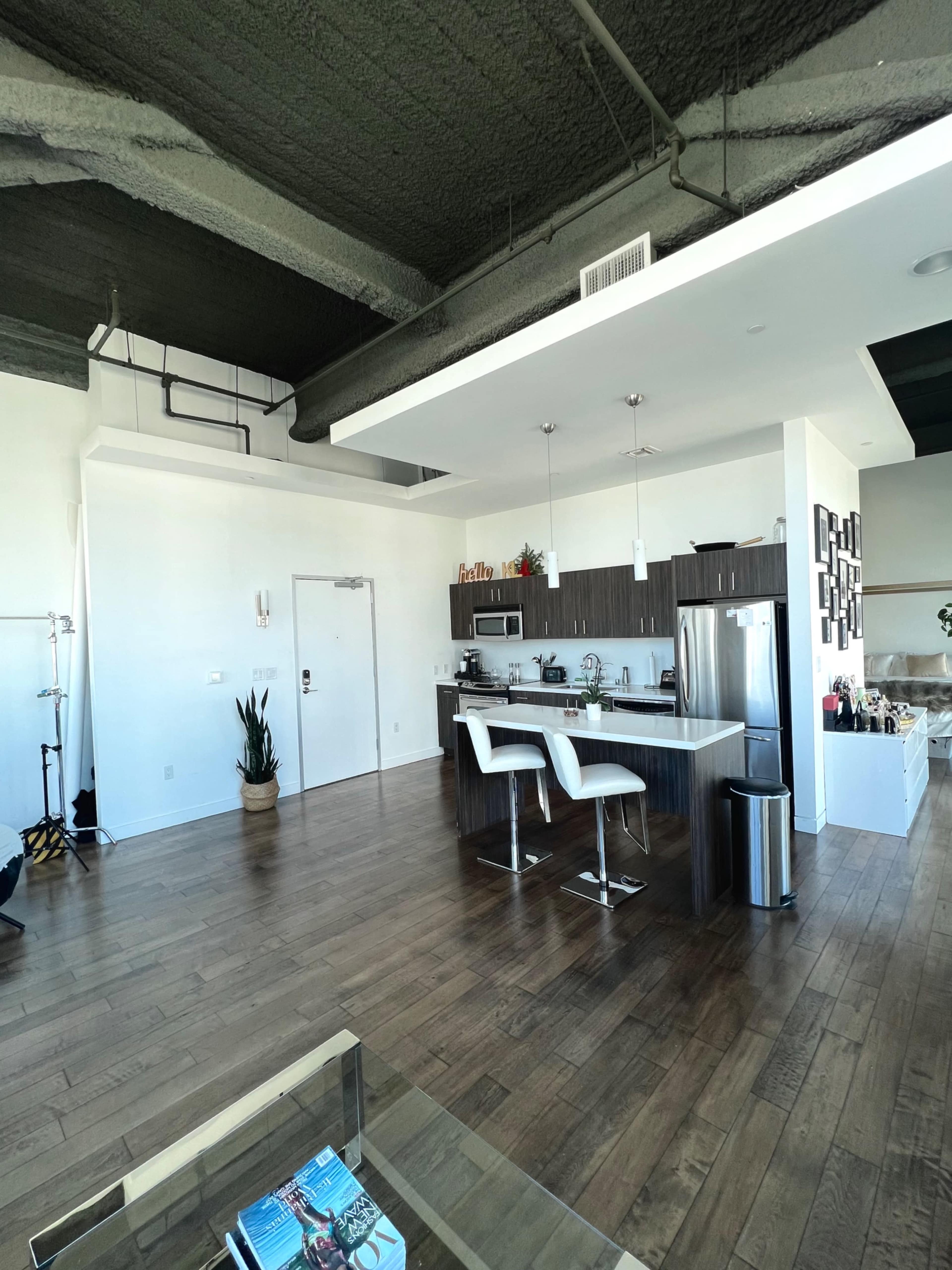 A modern kitchen with a bar area, featuring two white stools, dark cabinetry, and an open layout leading to a living space.