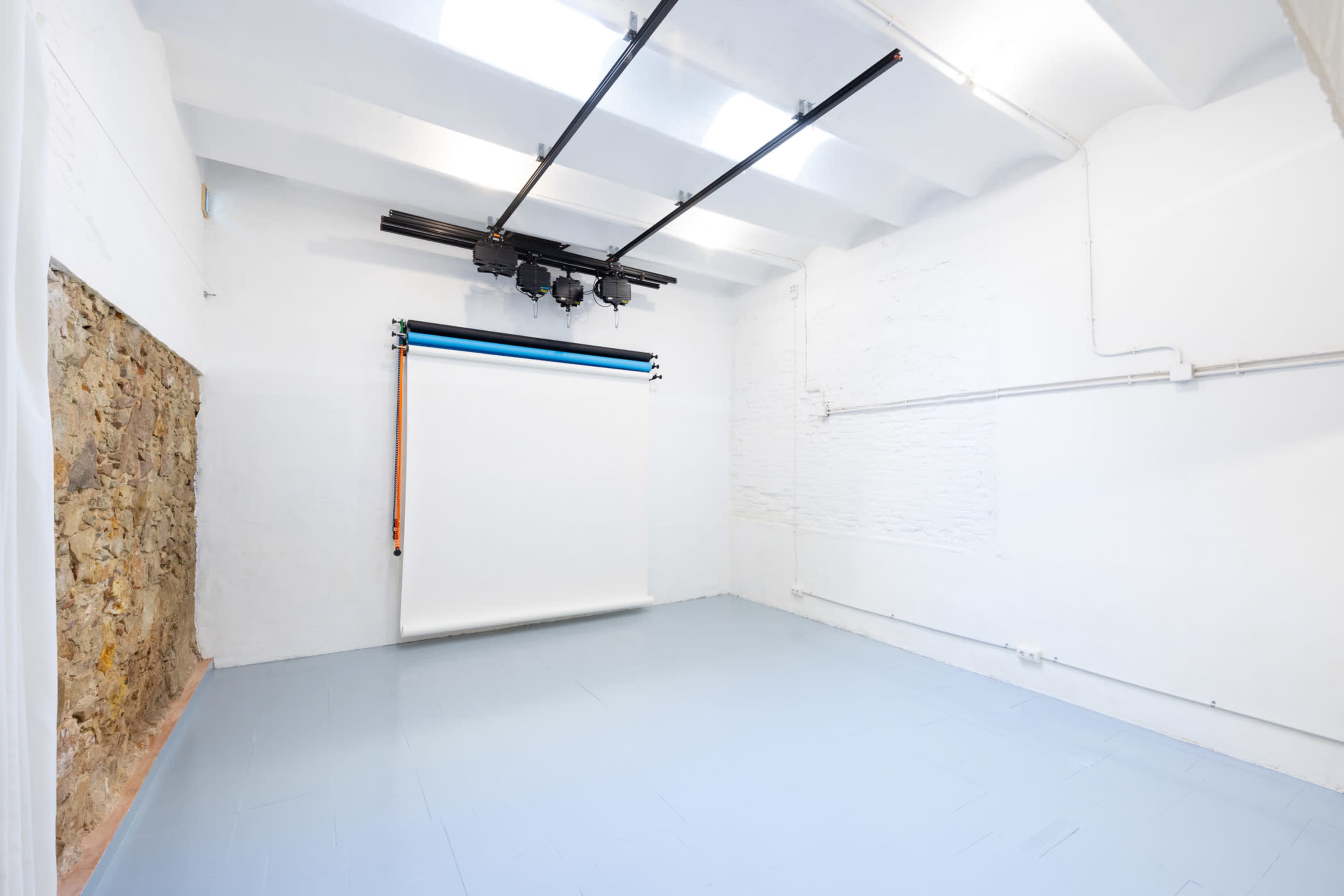 The image shows a spacious, well-lit studio with a smooth blue floor, a white backdrop hanging from a rail, and a section of exposed stone wall.