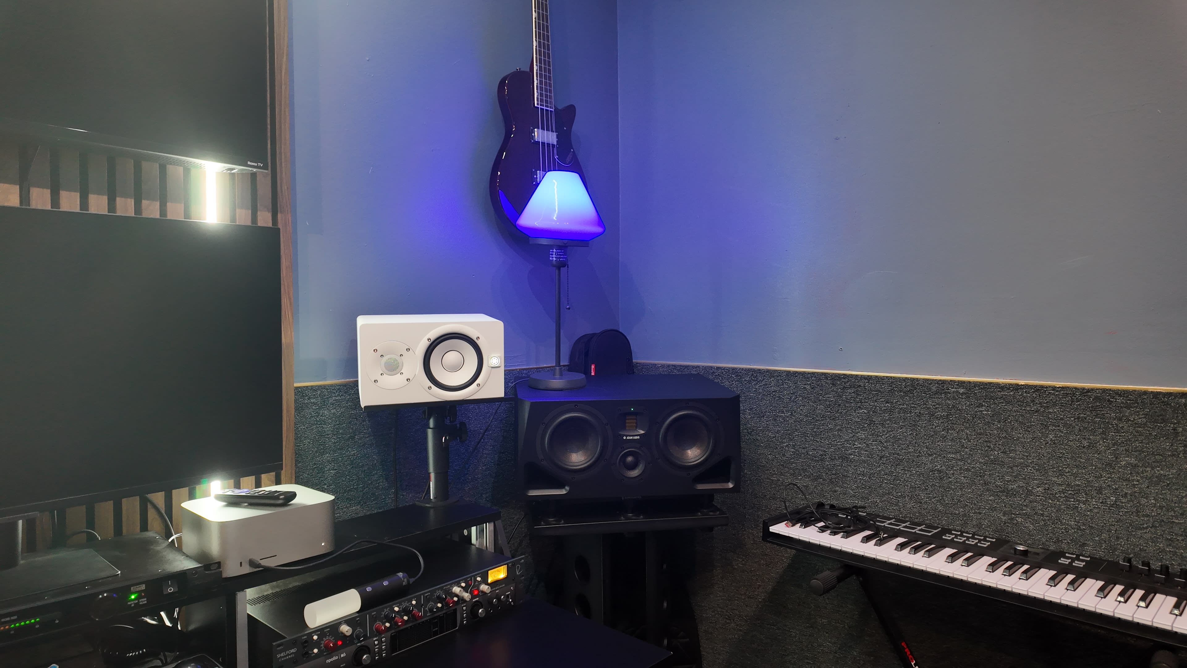 The image shows a music studio setup with a guitar mounted on the wall, a monitor speaker on a stand, a keyboard, and various audio equipment arranged along the walls.