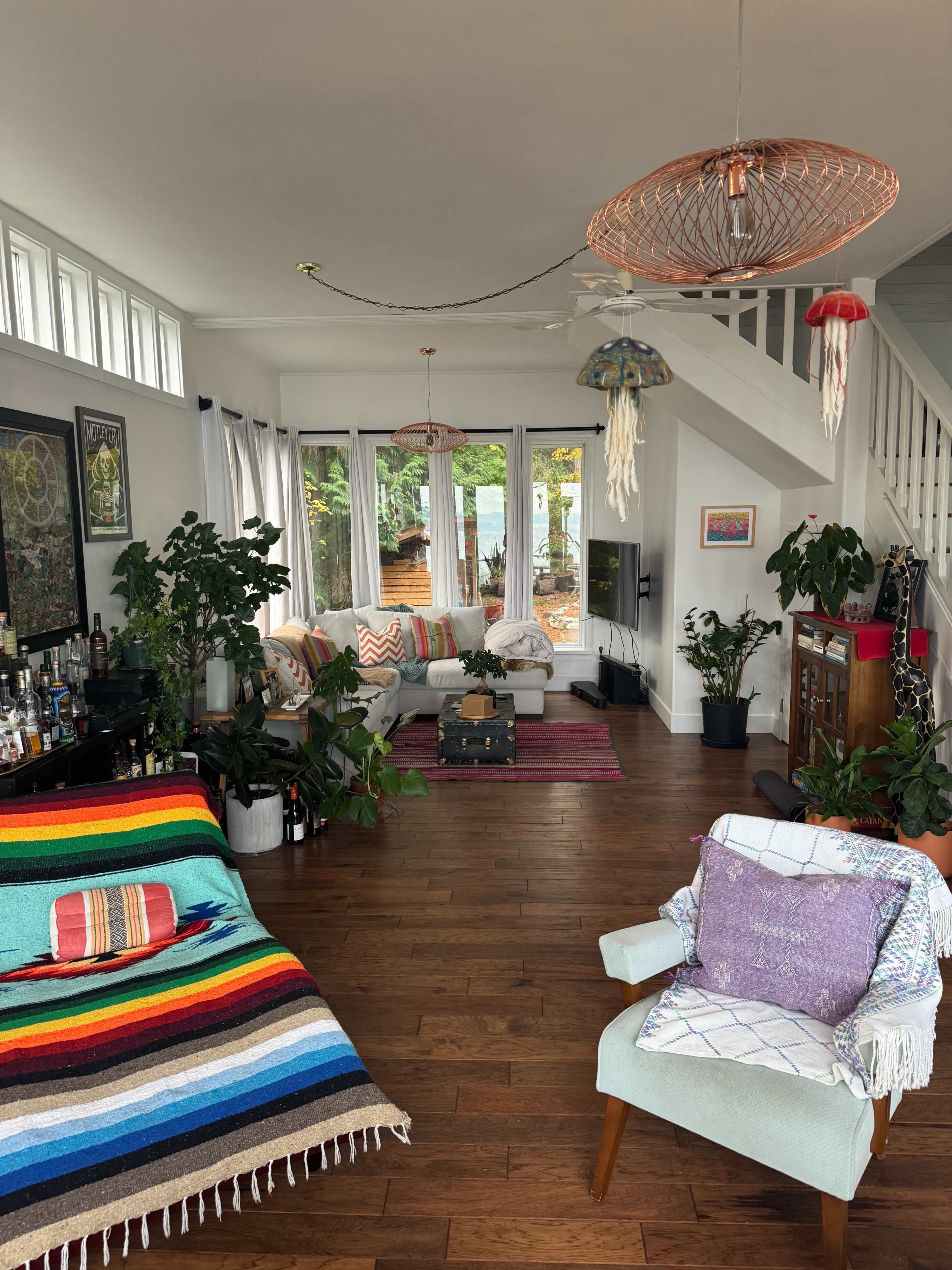 A living room features a striped blanket draped over a sofa, potted plants, and colorful pendant lights hanging from the ceiling.
