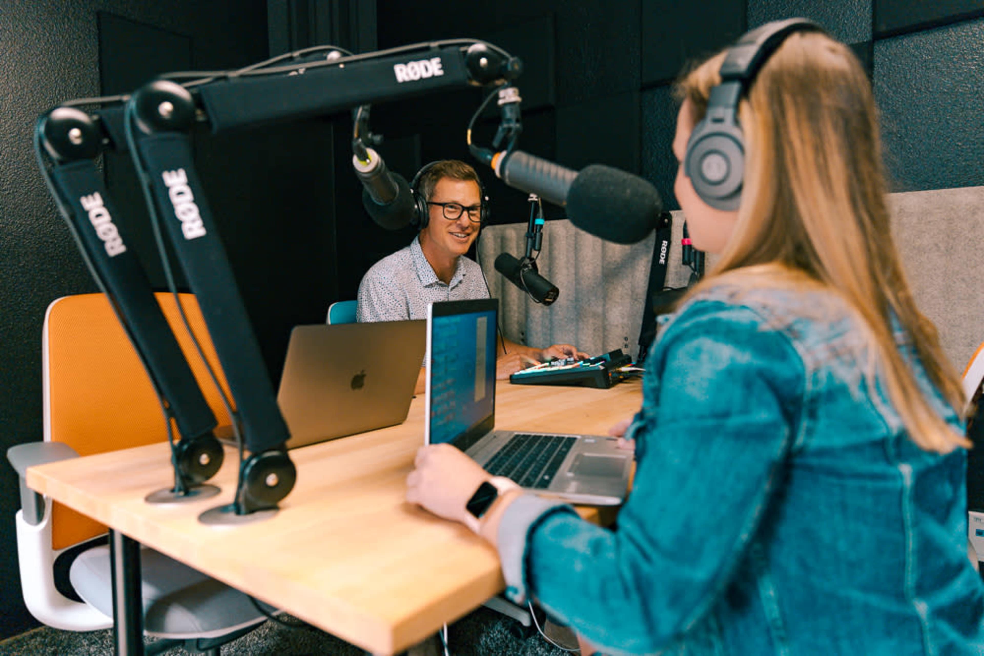A man and a woman sit at a table in a recording studio, each wearing headphones and using laptops while microphones are positioned in front of them.