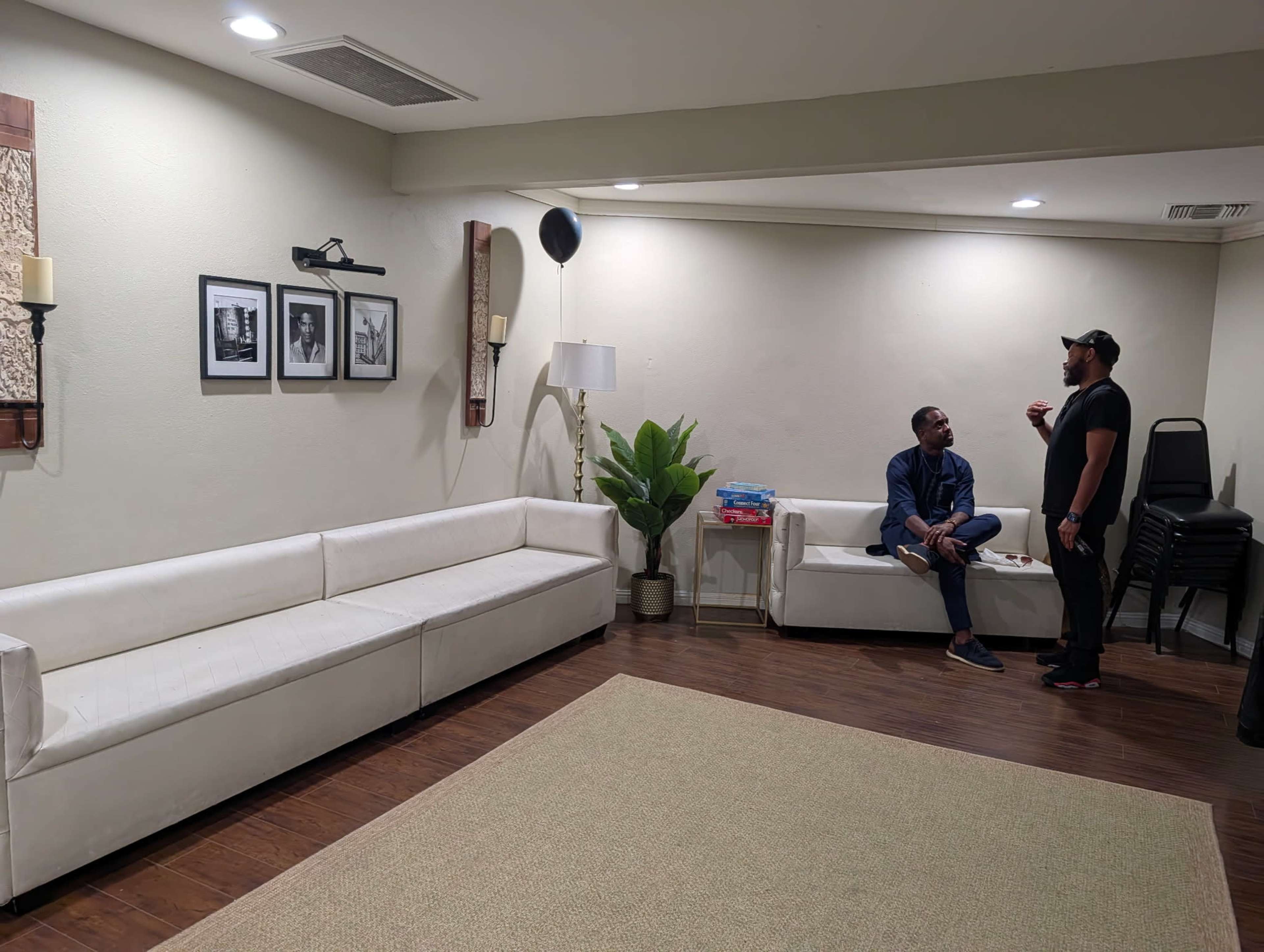 Two men are engaged in conversation in a well-lit room featuring white sofas, a decorative plant, and framed black-and-white photos on the wall.
