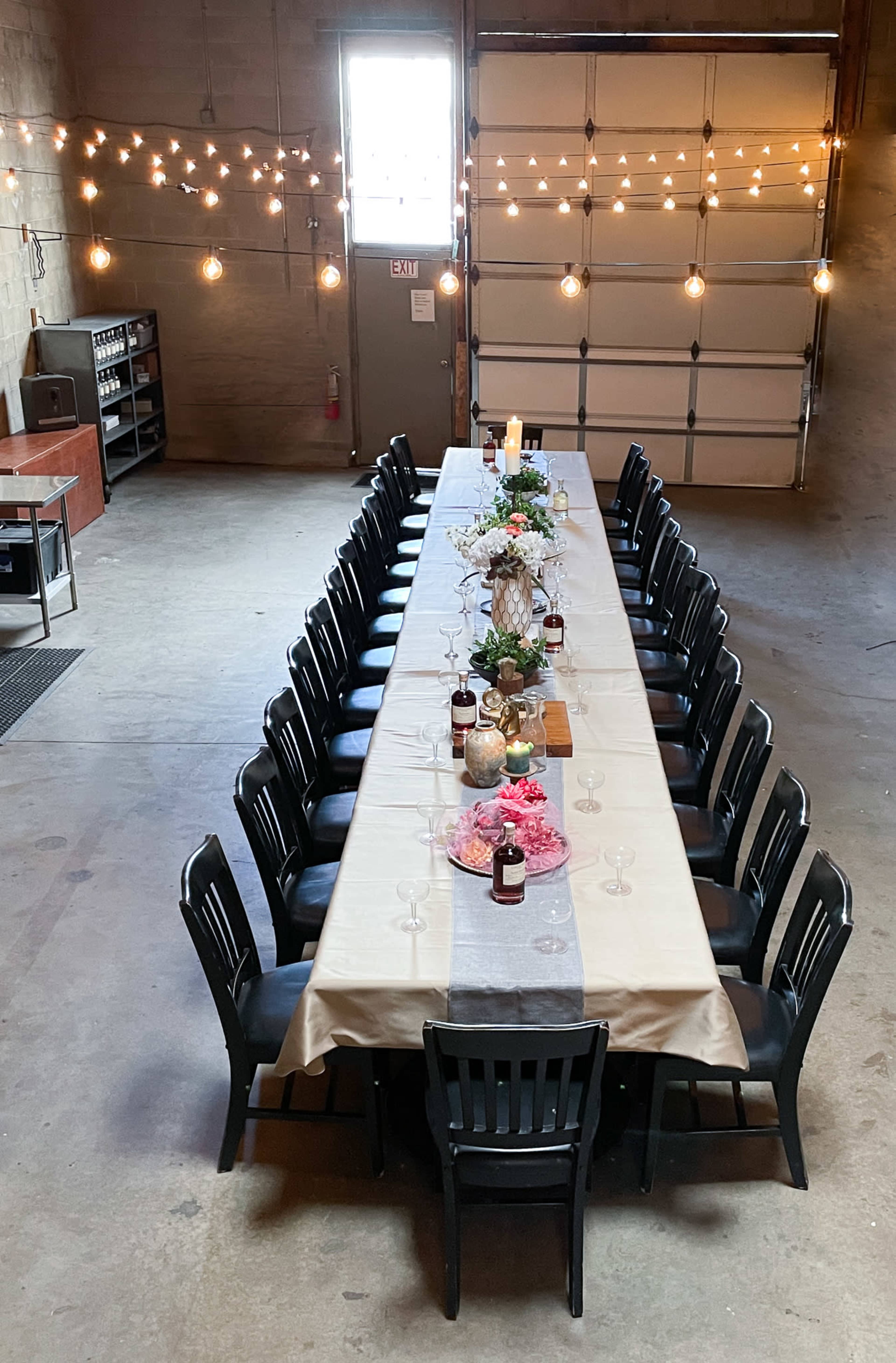 A long table with chairs is set up in a spacious garage, illuminated by string lights hanging from the ceiling.