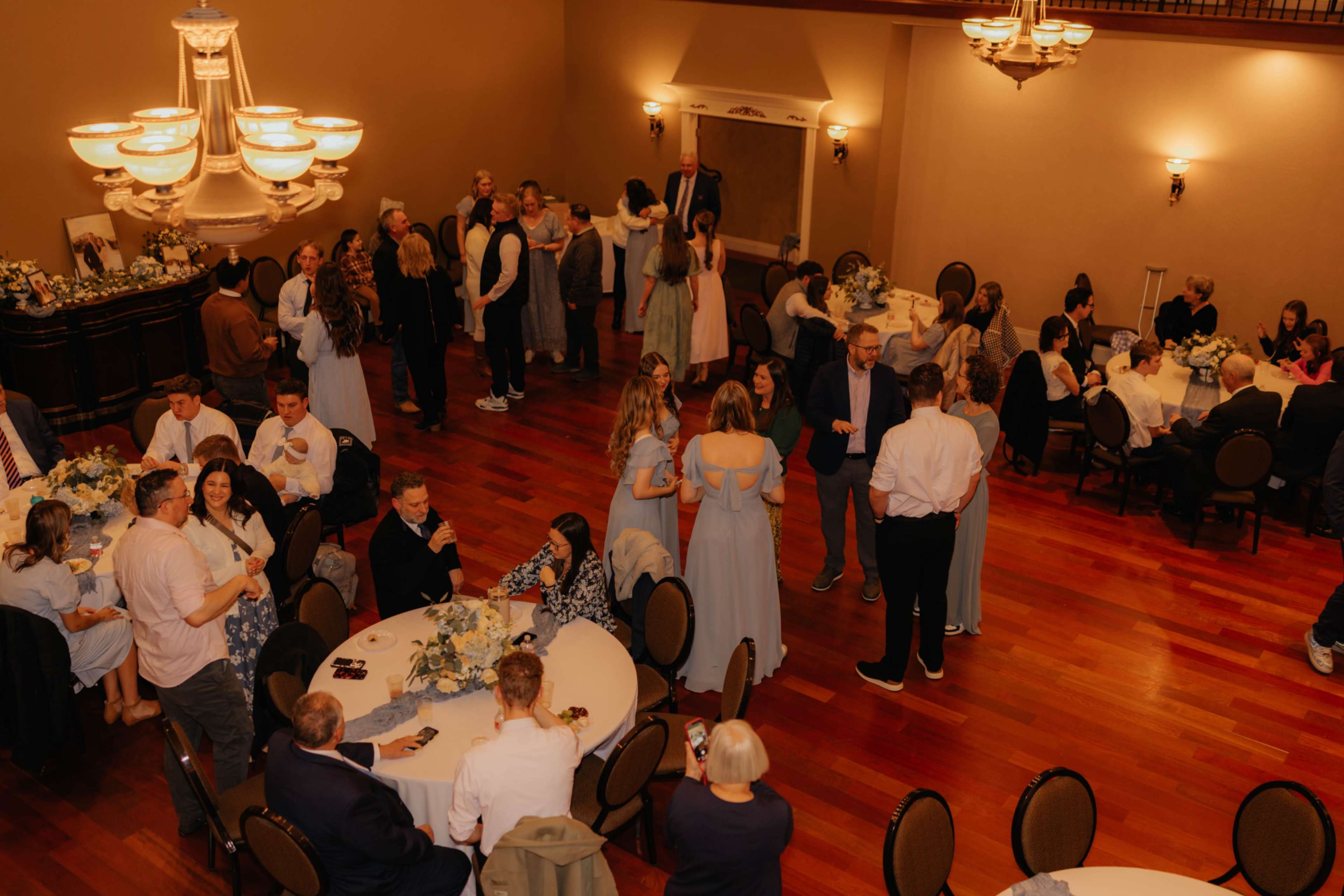 A large group of people is gathered in a banquet hall, engaged in conversation and celebrating around circular tables decorated with flowers.