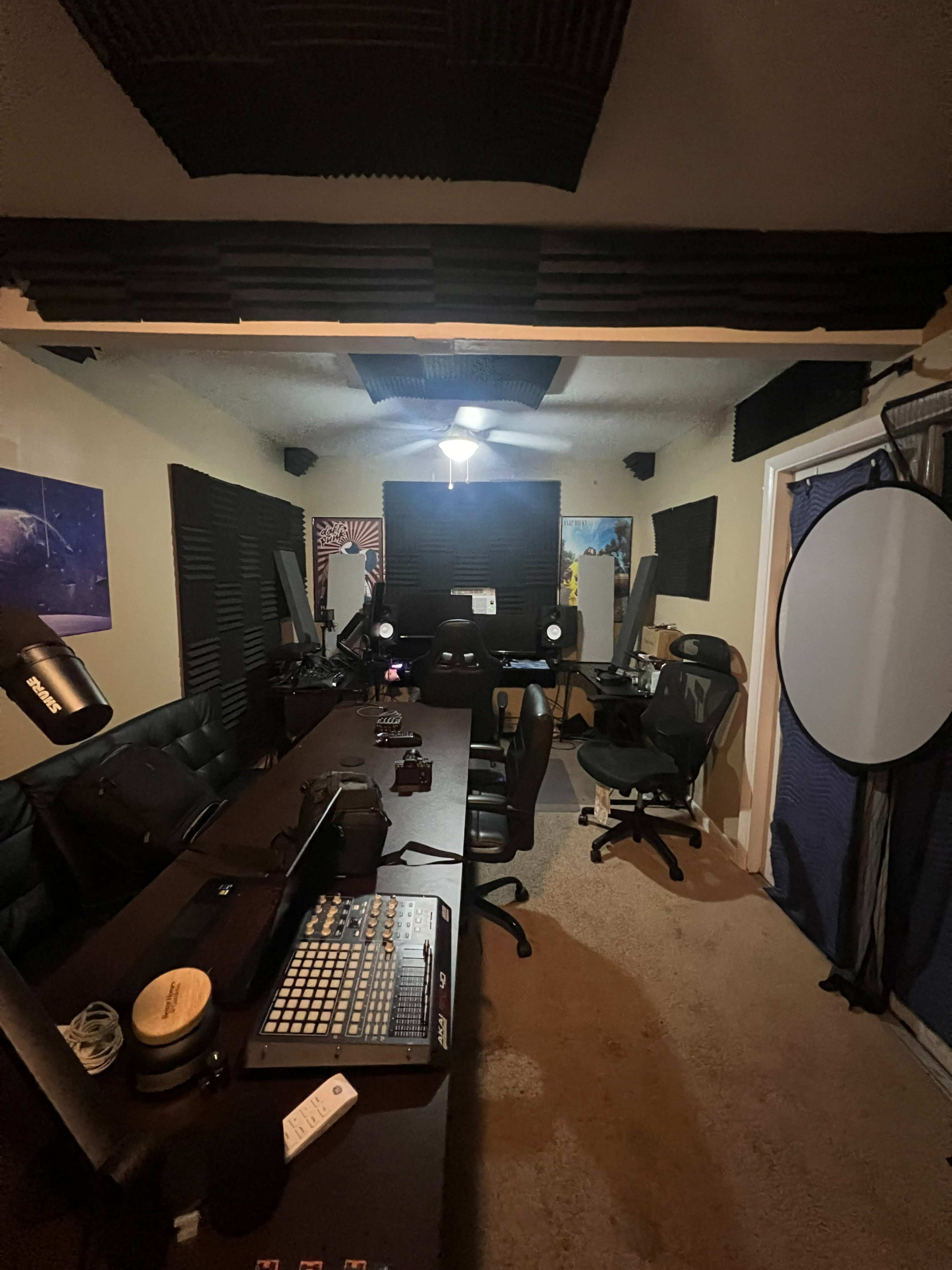 A music production studio featuring a long table with equipment, soundproofing panels on the walls, and various chairs arranged in the space.