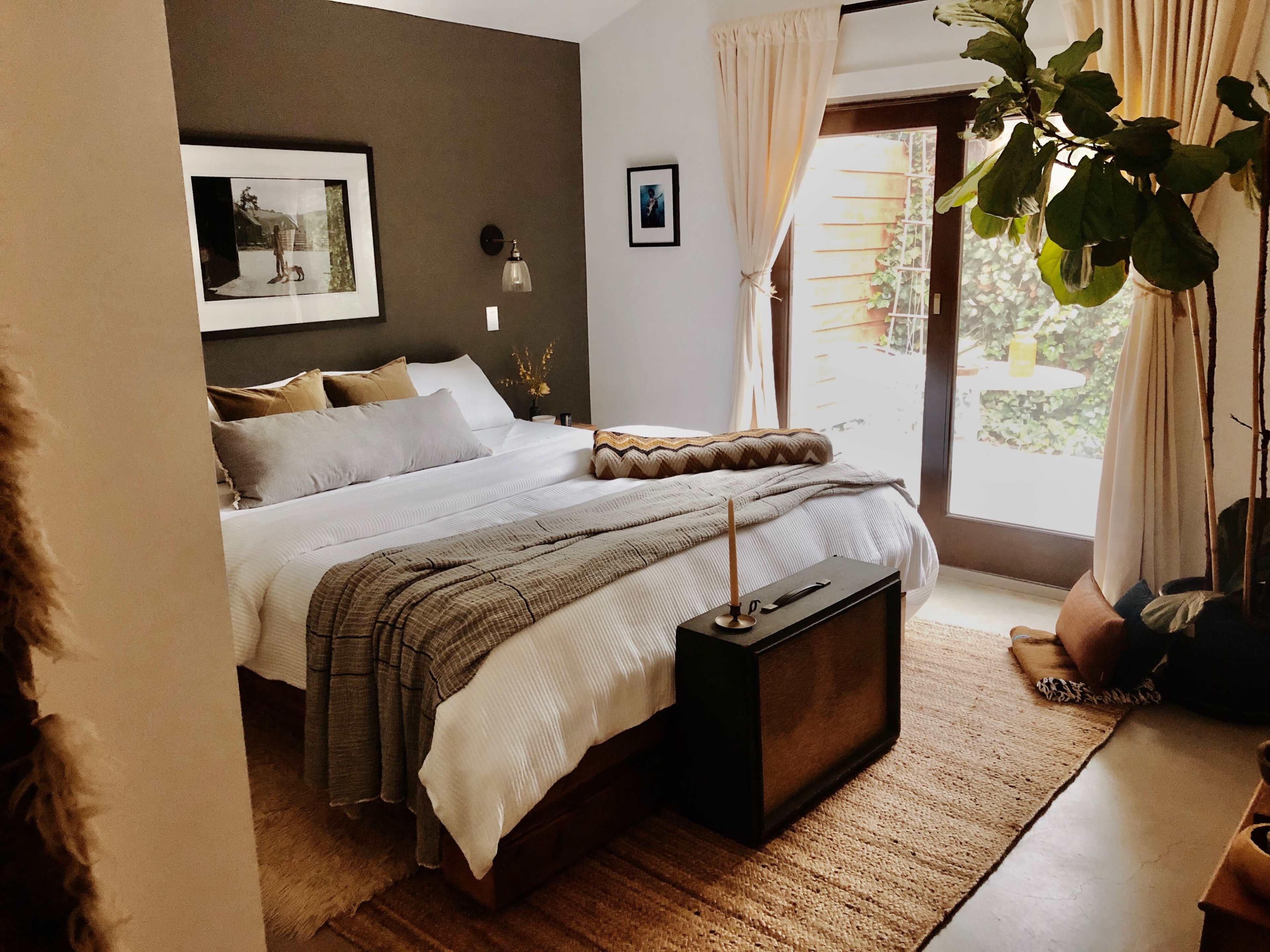 A minimalist bedroom features a king-sized bed with neutral bedding, a nightstand with a candle, and a large window opening to a garden view.