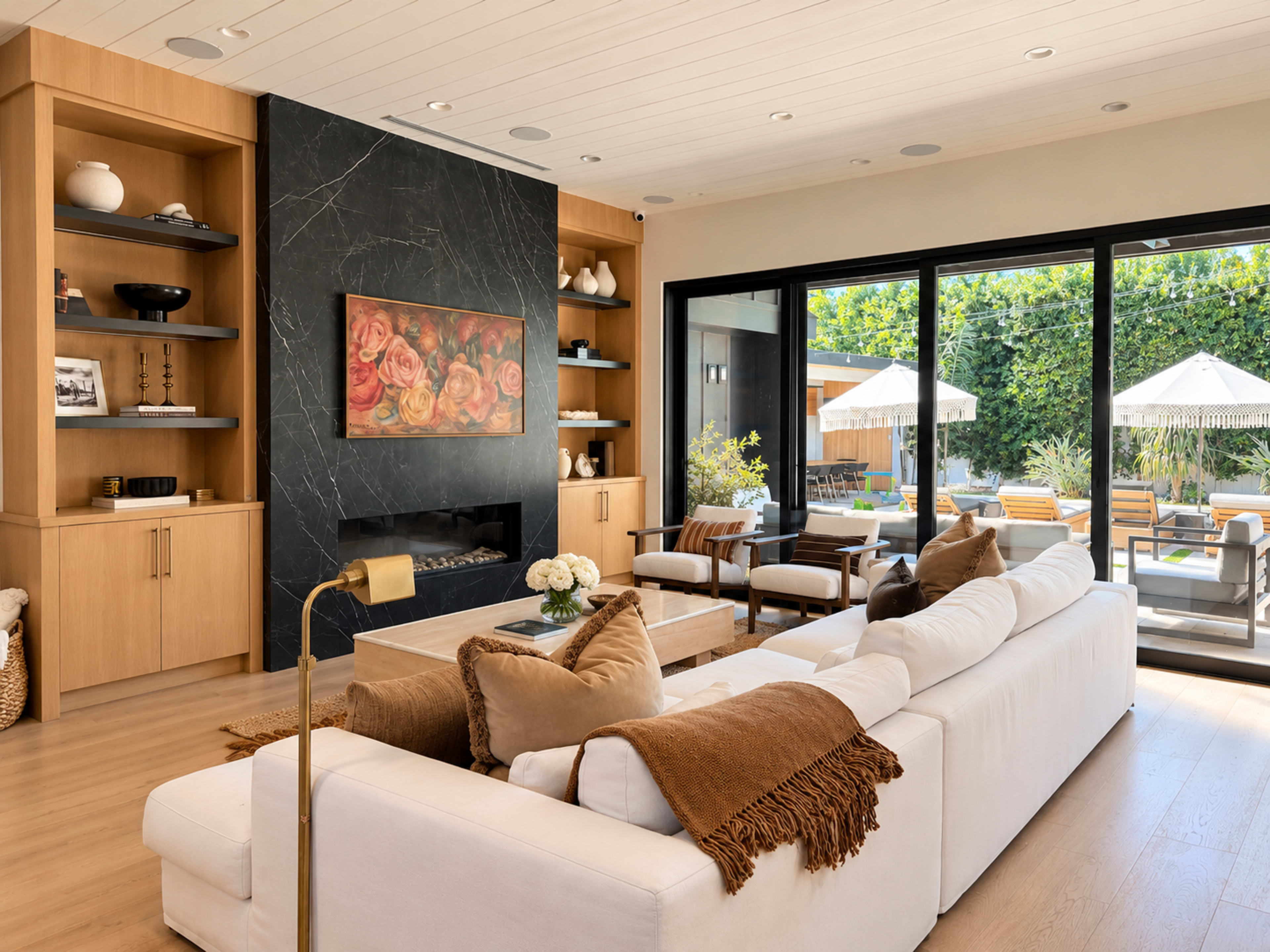 A modern living room features a white sofa with brown throw pillows, a marble accent wall with a floral painting, and large glass doors that open to a patio with outdoor seating.