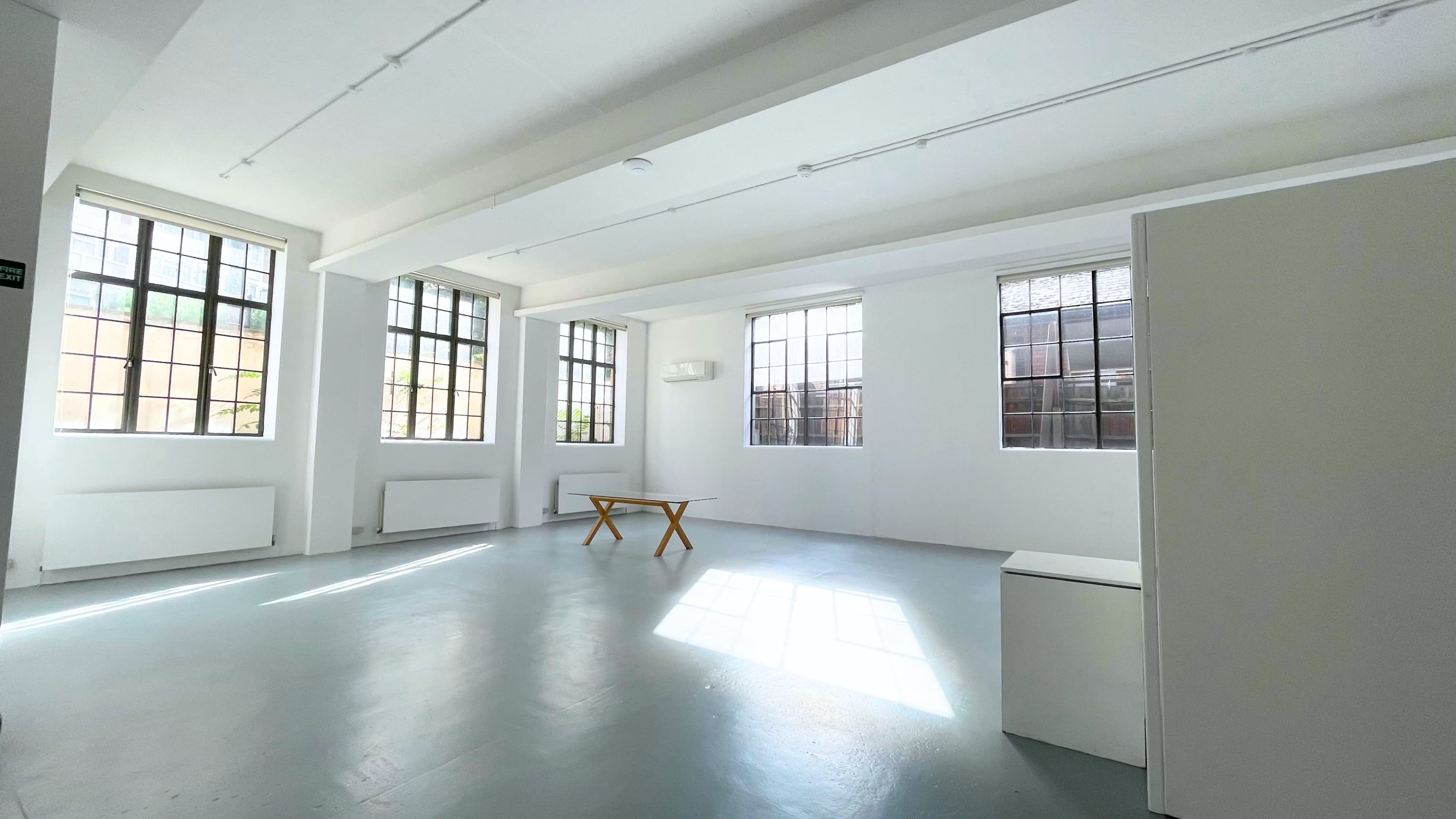 A spacious, well-lit room with large windows, a wooden table in the center, and minimal furnishings.