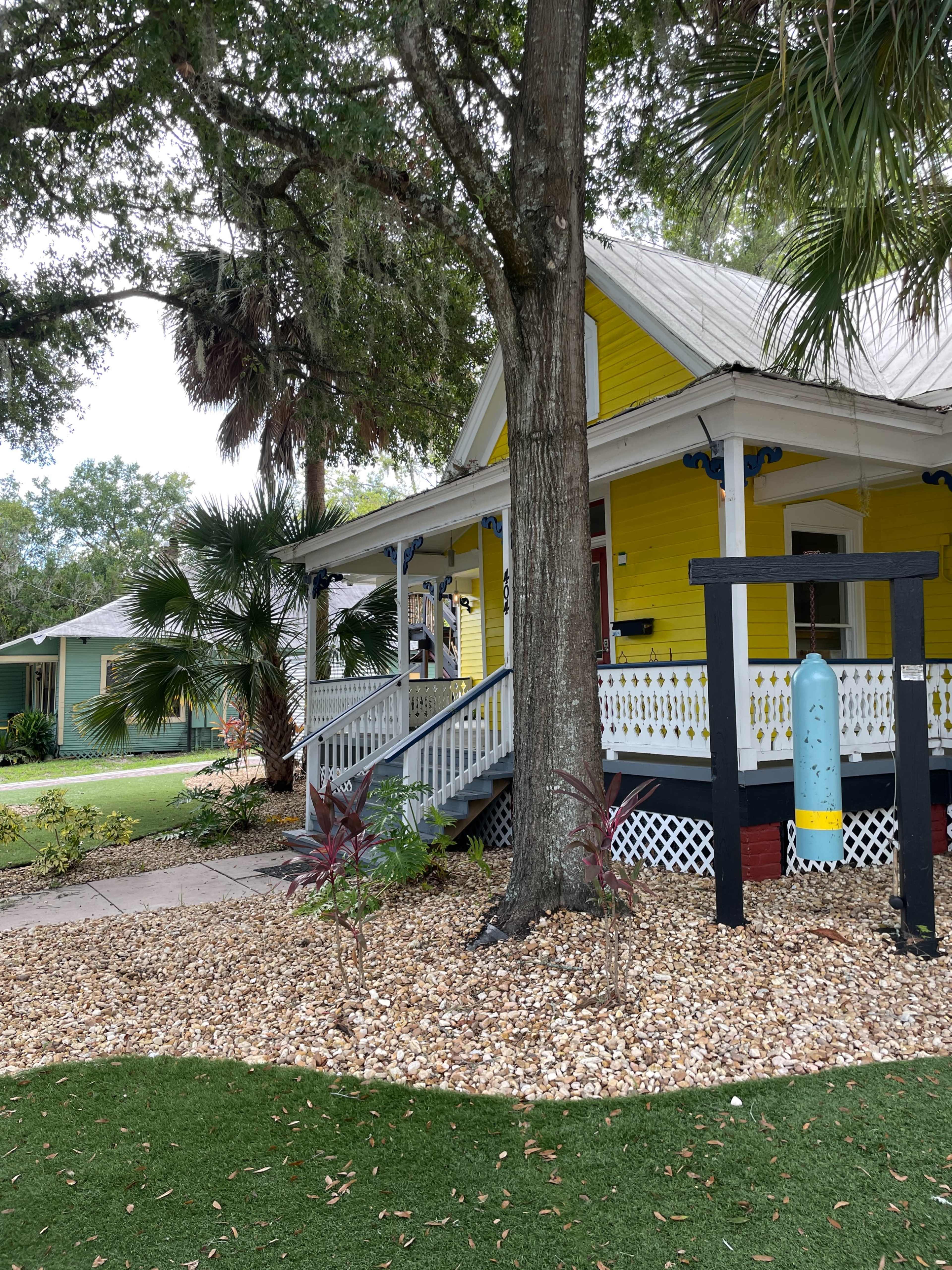 A yellow house with a porch is situated next to a large tree and surrounded by decorative landscaping made of small rocks and plants.