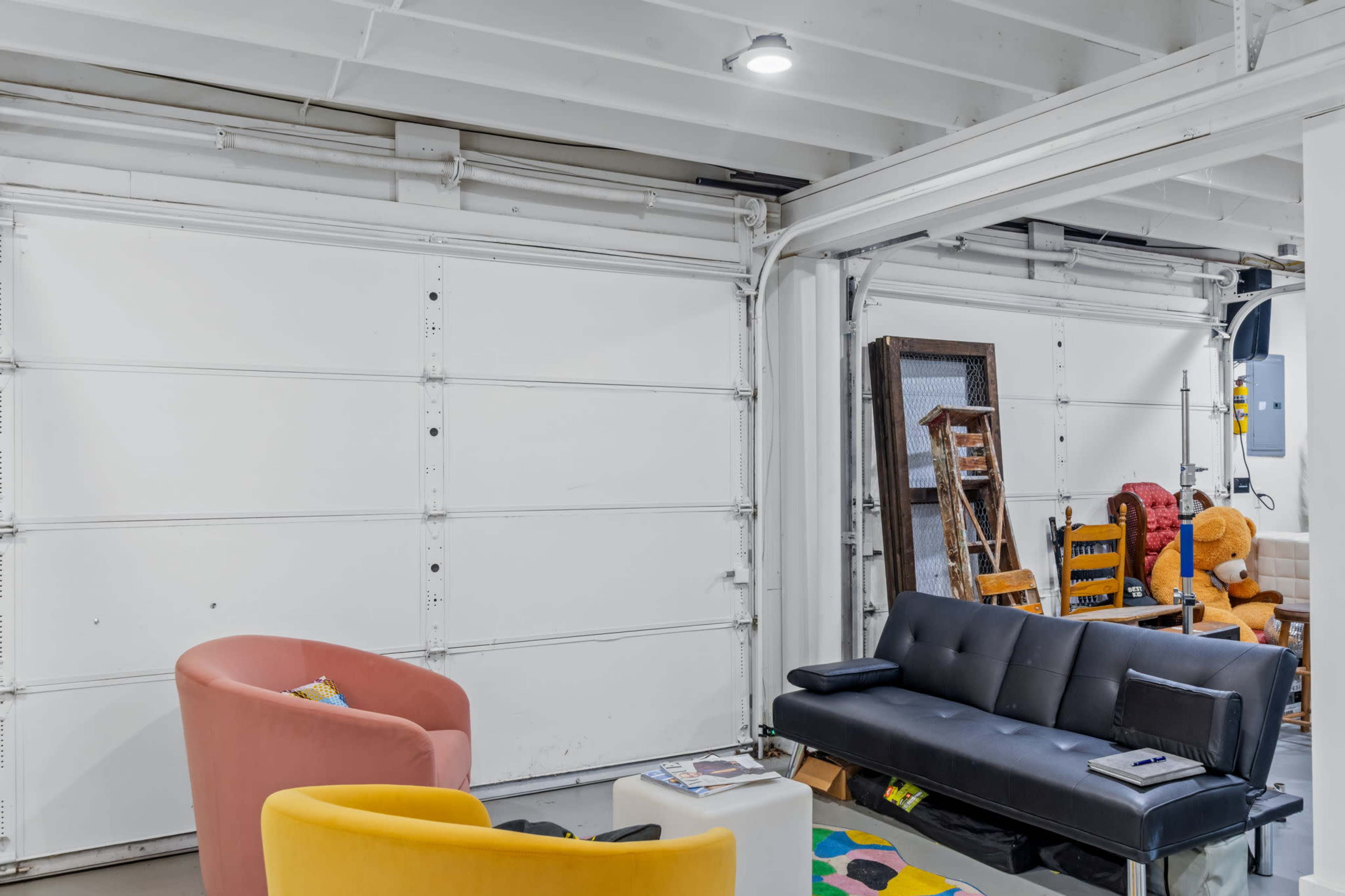 The image shows a modern garage space converted into a living area, featuring a pink chair, a yellow chair, and a black sofa, with a white garage door in the background.