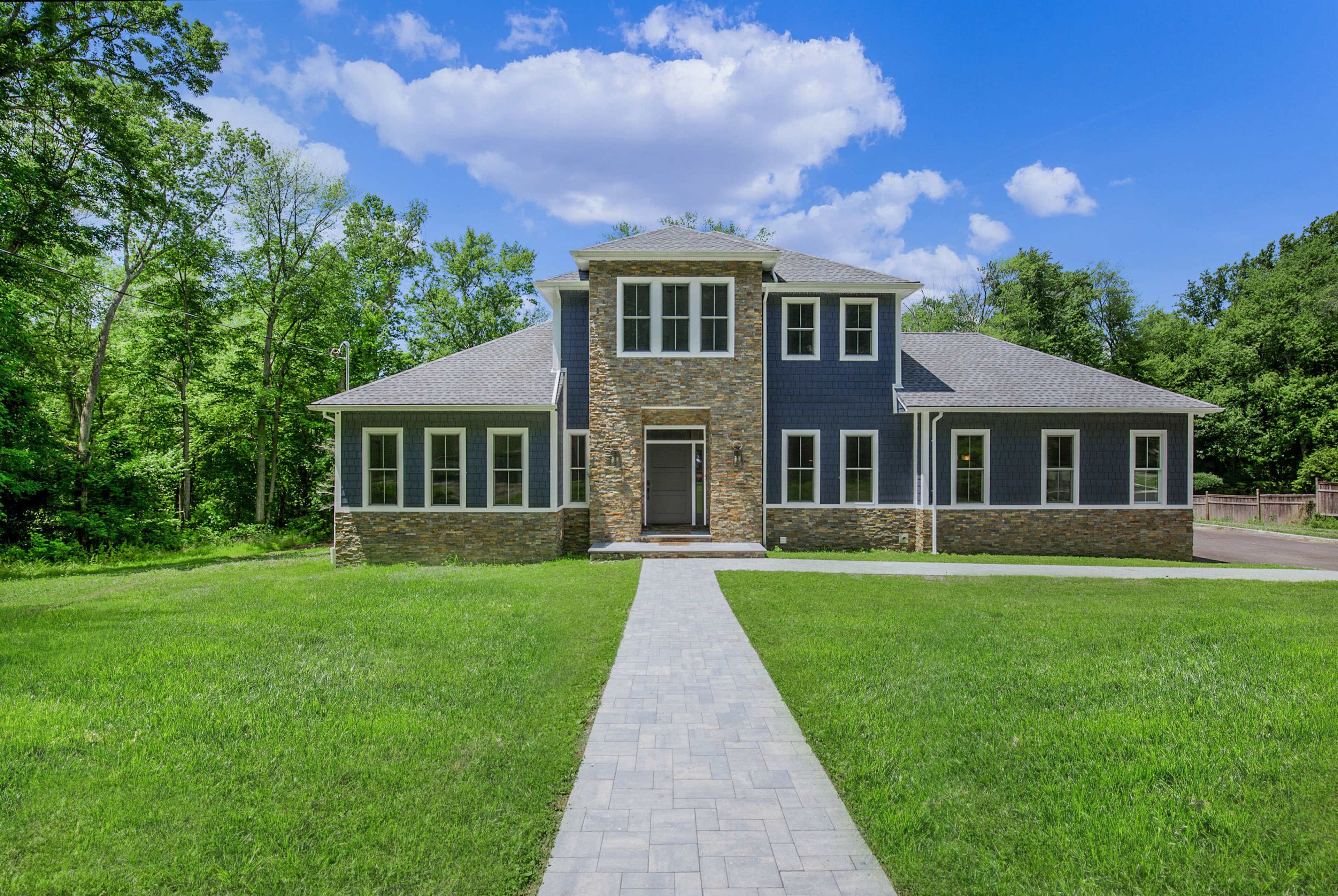A large, modern two-story house with a stone and wood exterior, surrounded by a grassy area and trees under a blue sky.