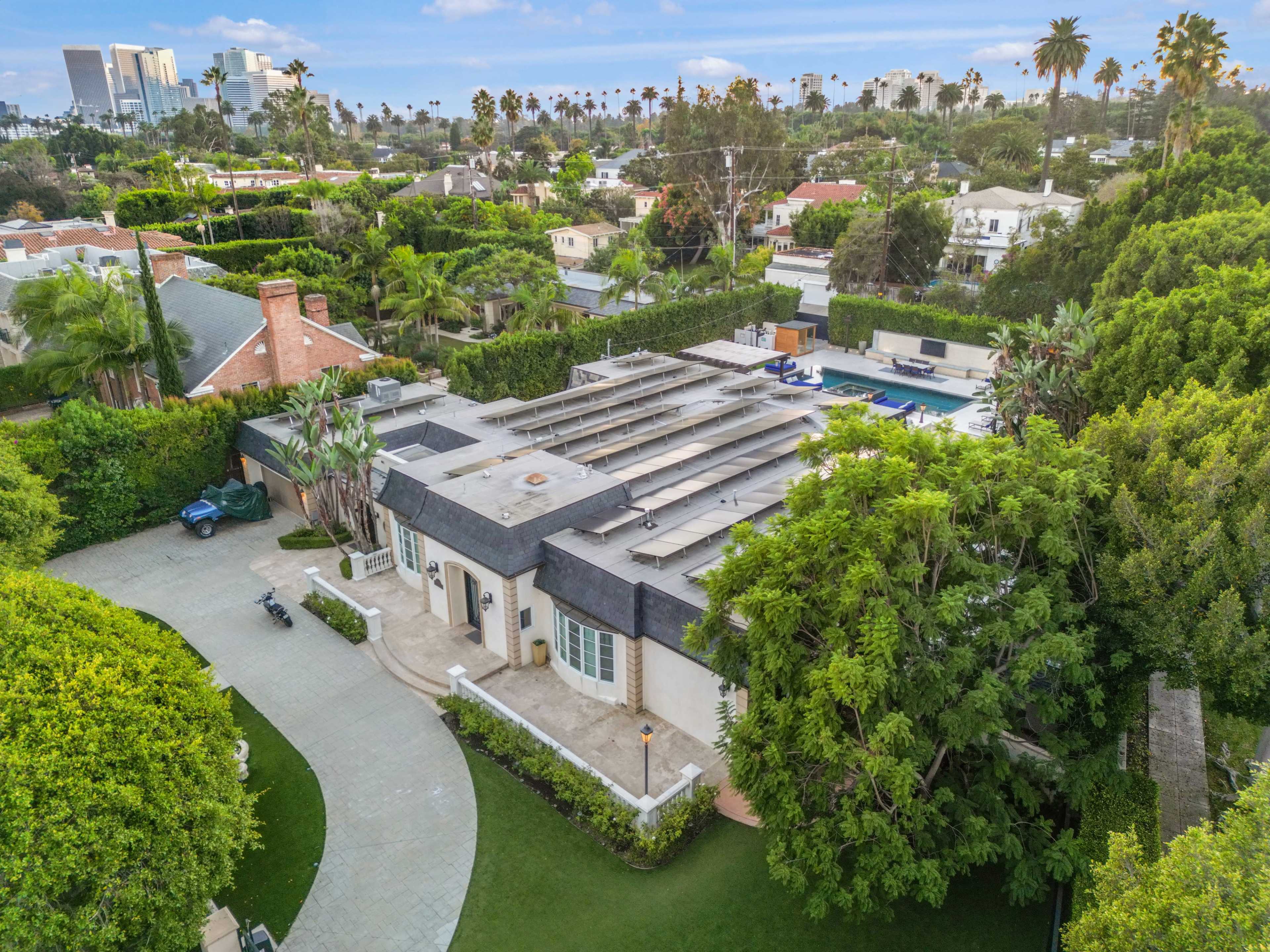 The image shows a large, modern home surrounded by lush greenery and trees, with a swimming pool visible in the backyard and city buildings in the background.