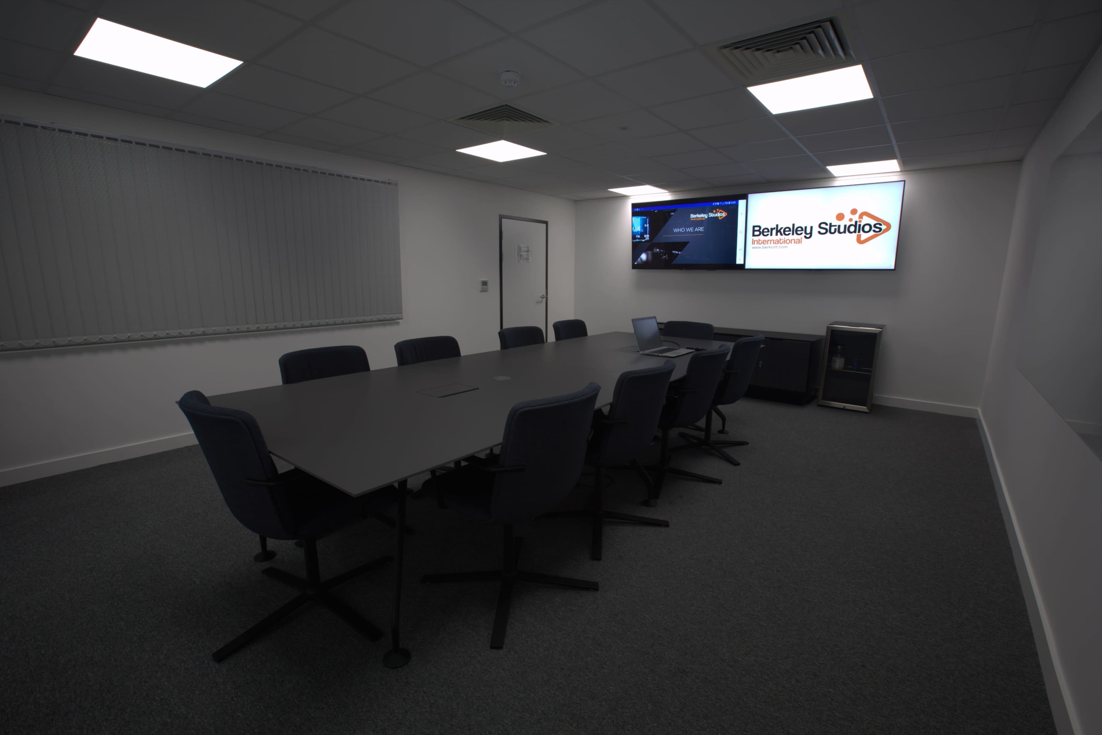 The image shows a modern conference room with a long table surrounded by chairs, equipped with a large screen on one wall.
