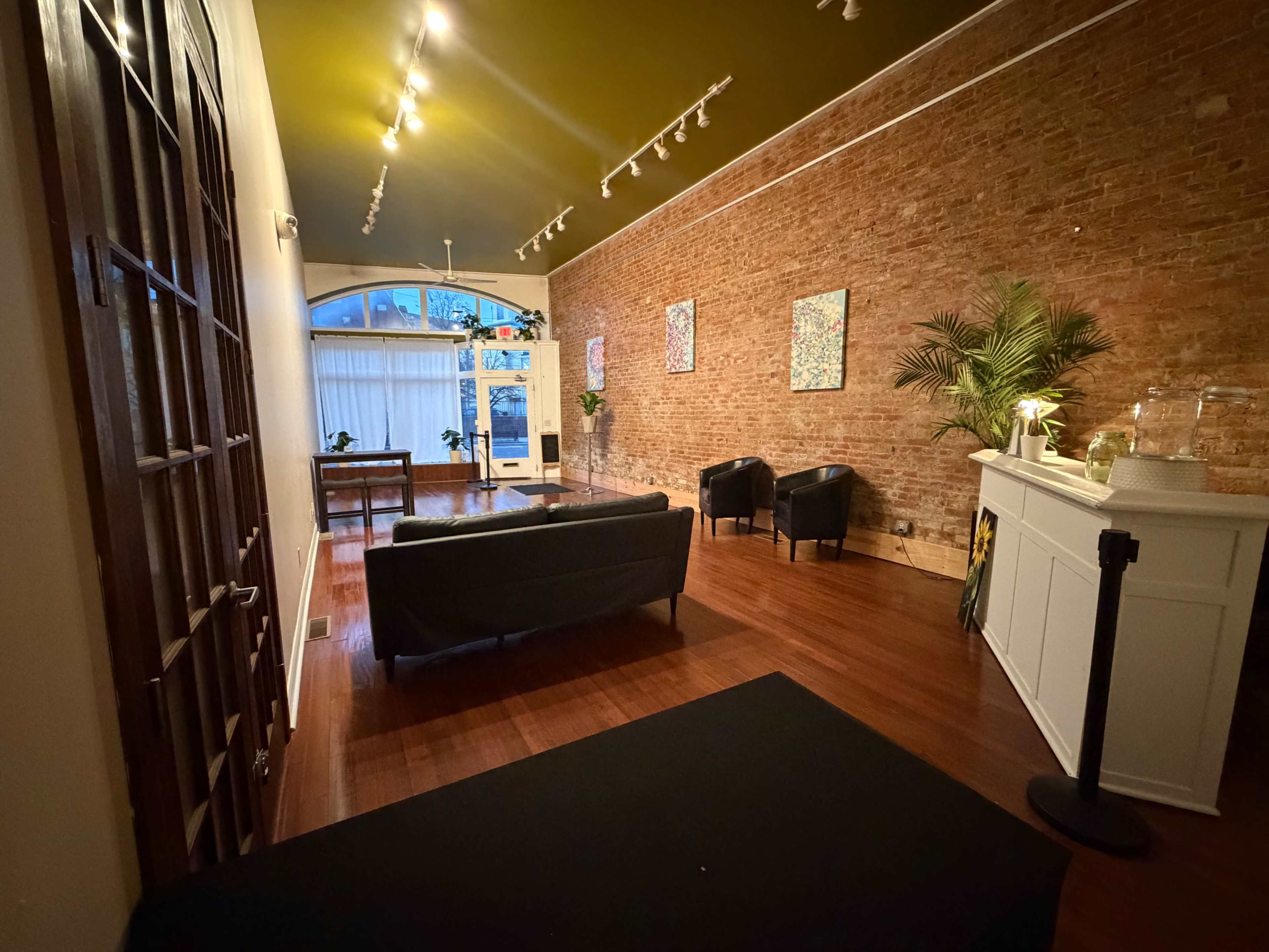 The image shows a spacious, well-lit interior featuring a wooden floor, exposed brick walls, and modern furnishings, including black sofas and decorative art on the walls.