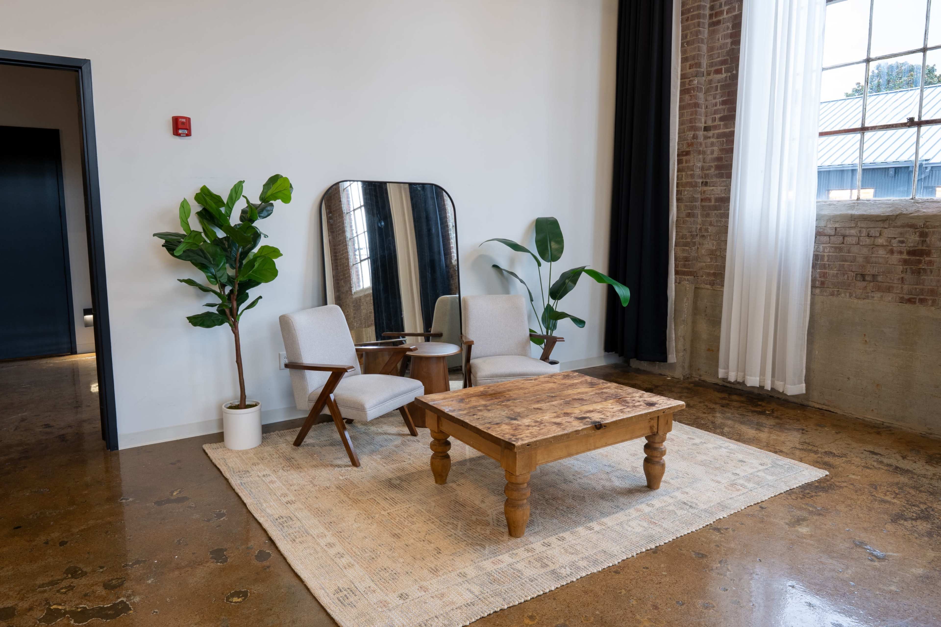 The scene features two chairs positioned beside a wooden coffee table on a rug, with a large mirror and potted plants against a backdrop of exposed brick walls and large windows.