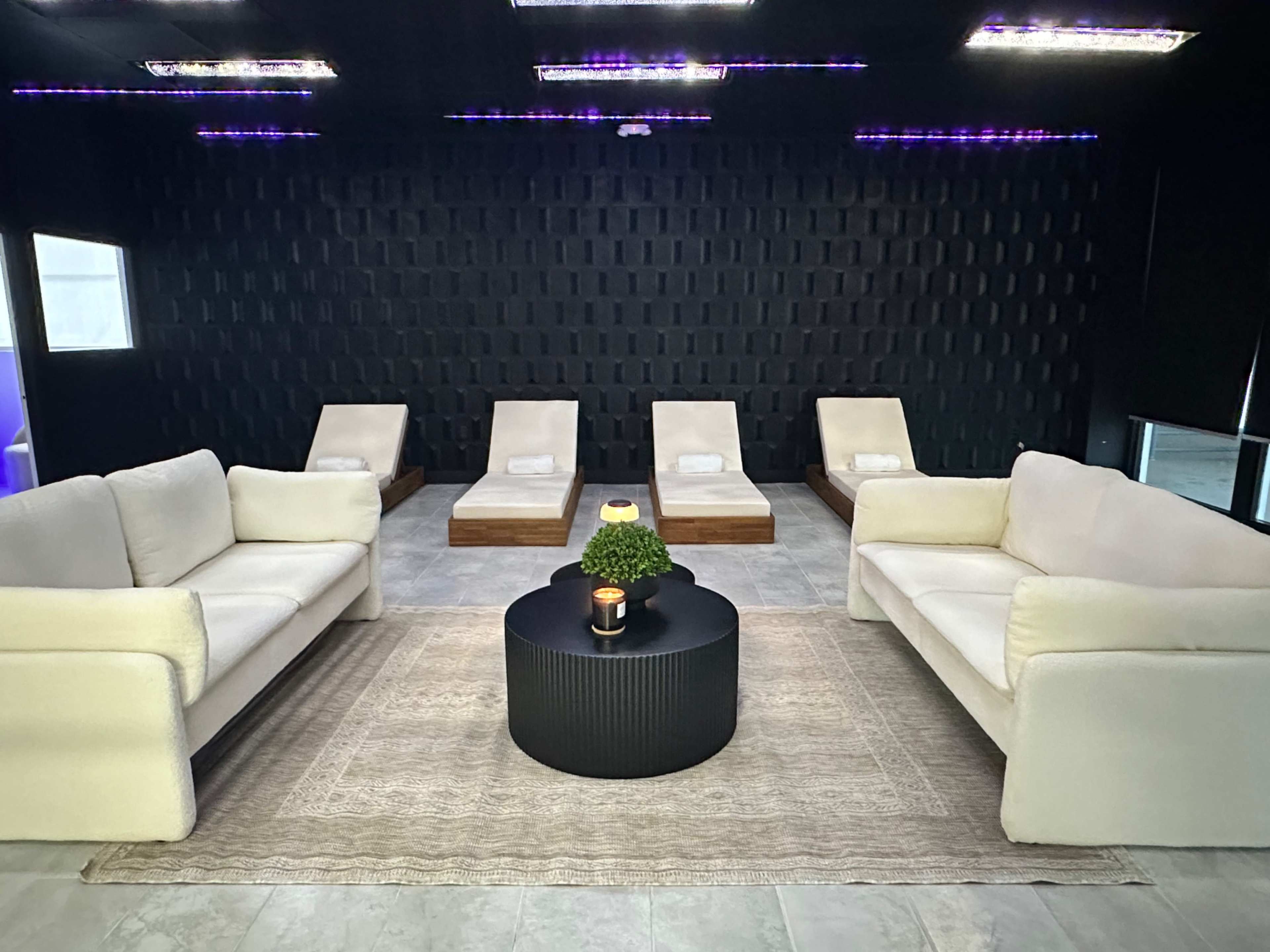 A dark room with white sofas and loungers arranged around a central black coffee table, featuring a small plant and candle.