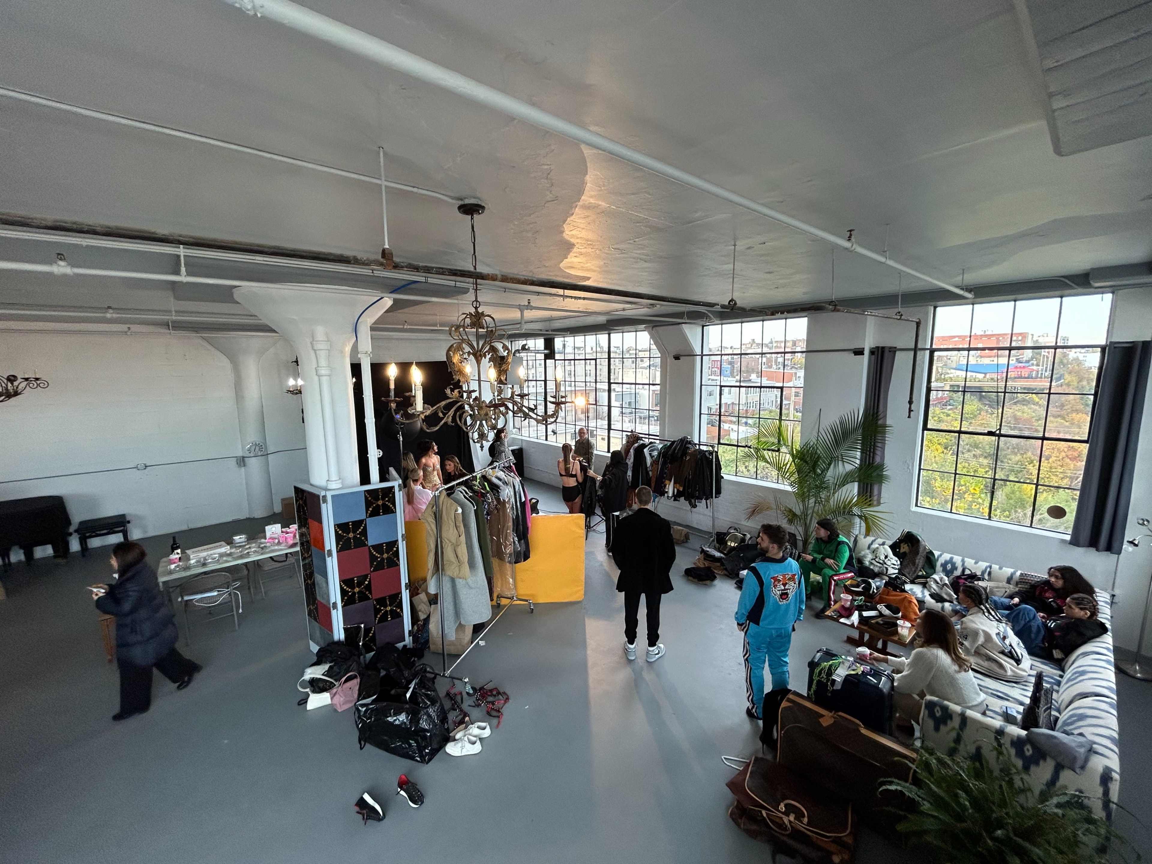 A spacious industrial-style room features various people socializing, clothing racks, and an array of bags and accessories scattered on the floor.