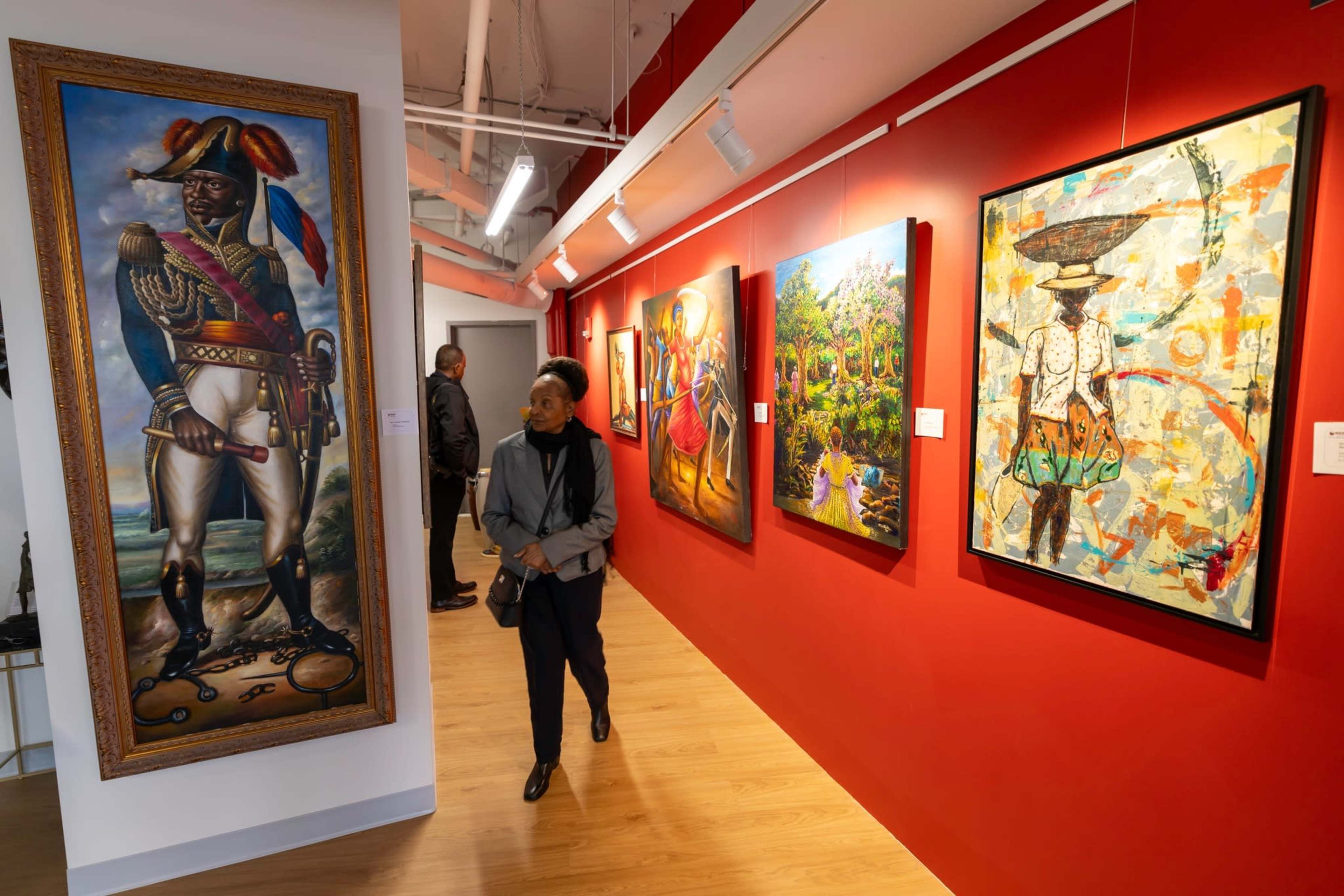 A person walks past colorful artworks displayed on red walls in a gallery, featuring paintings with diverse themes and styles.