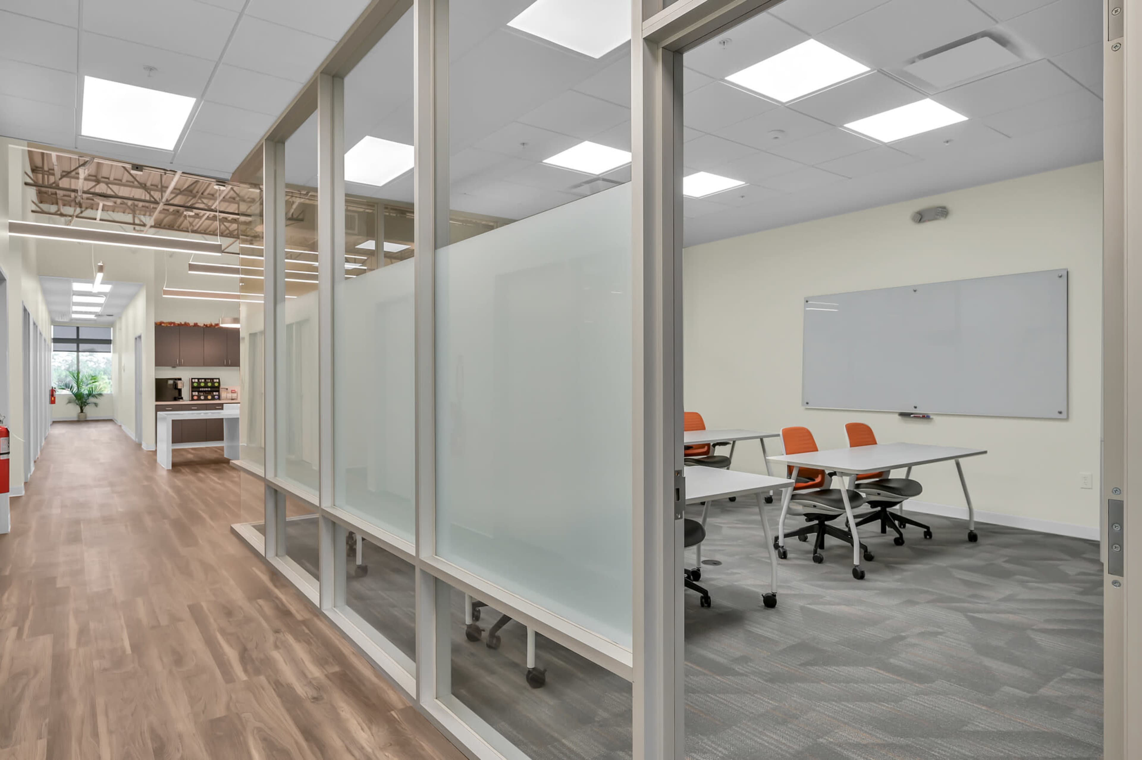 A modern office space with glass-paneled meeting rooms, orange chairs, and a hallway leading to a well-lit area with a whiteboard.