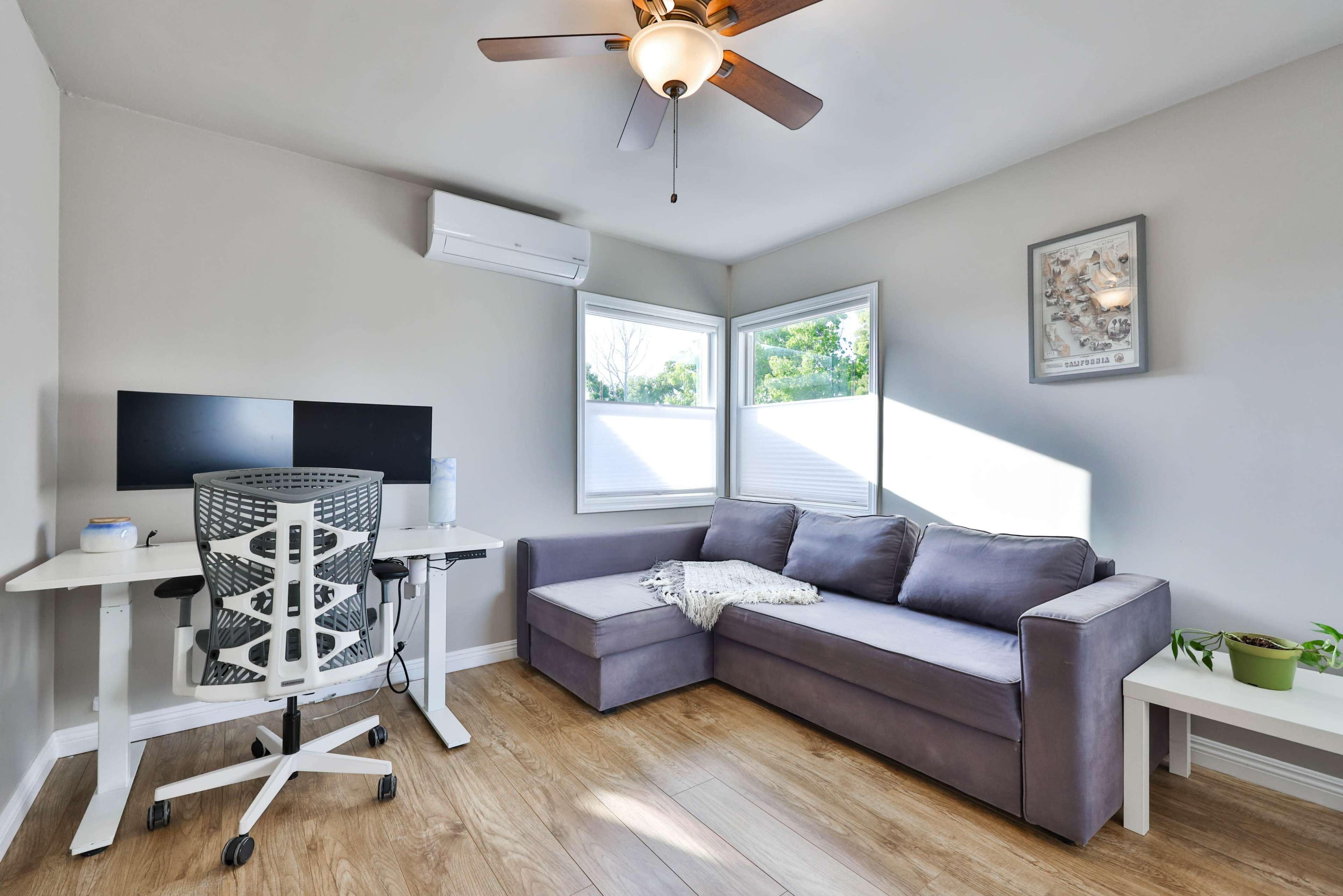 A modern, well-lit room featuring a gray sofa, a minimalist desk with dual monitors, and large windows that provide natural light.