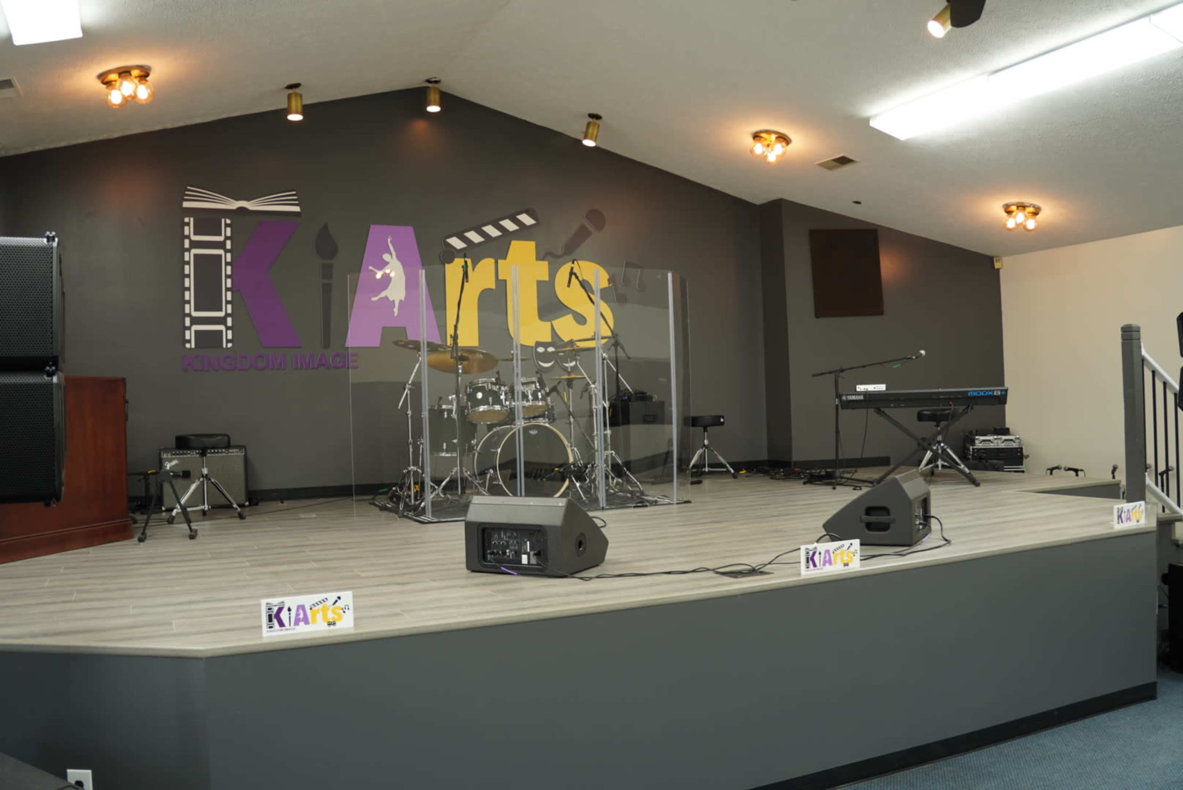 The image shows a stage set up for a performance, featuring a drum kit, keyboard, and sound equipment, with a large "KI Arts" sign on the wall behind it.