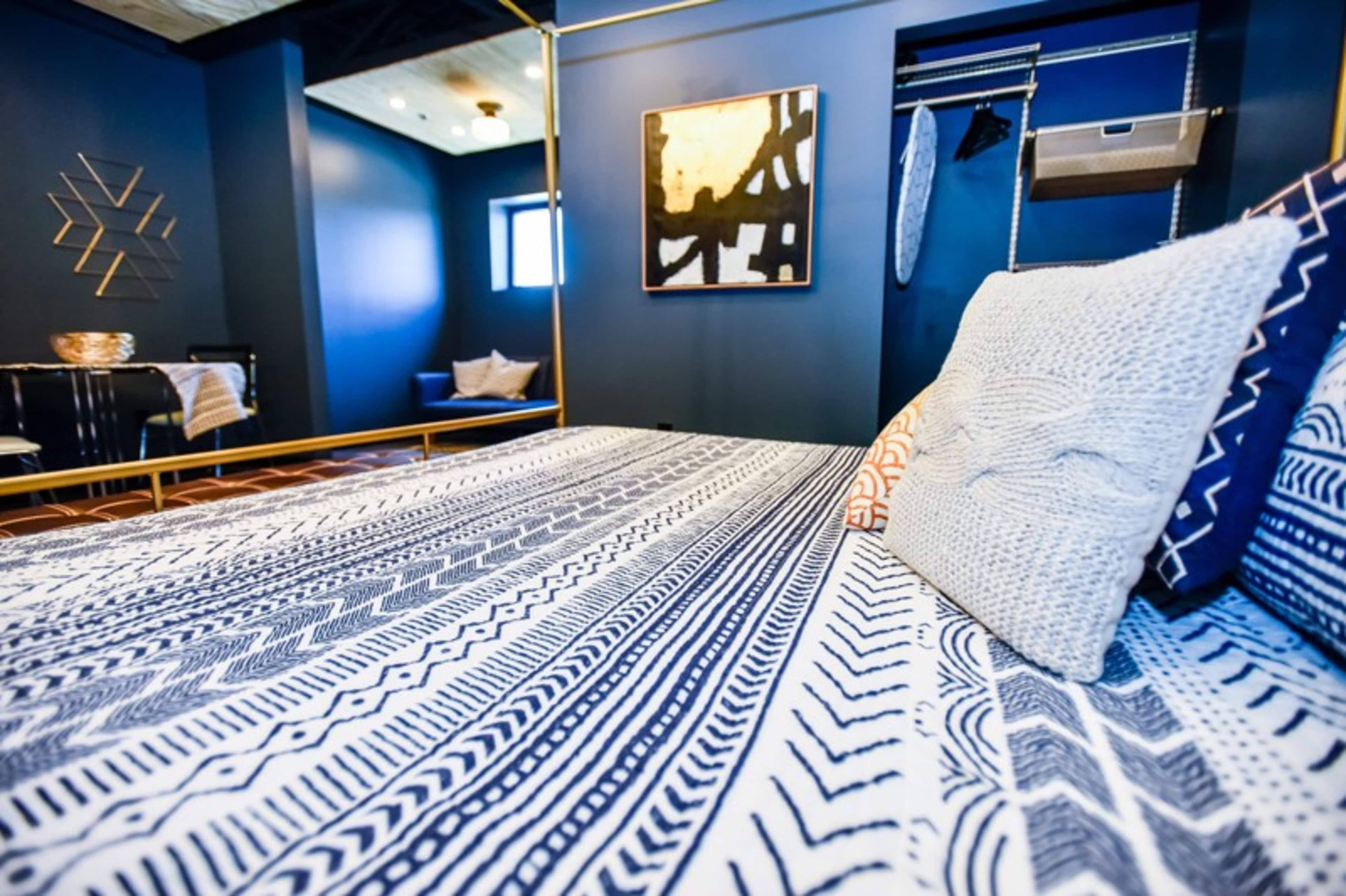 The image shows a modern bedroom featuring a patterned bedspread, a blue accent wall, and decorative elements like a framed artwork and a clothing rack.