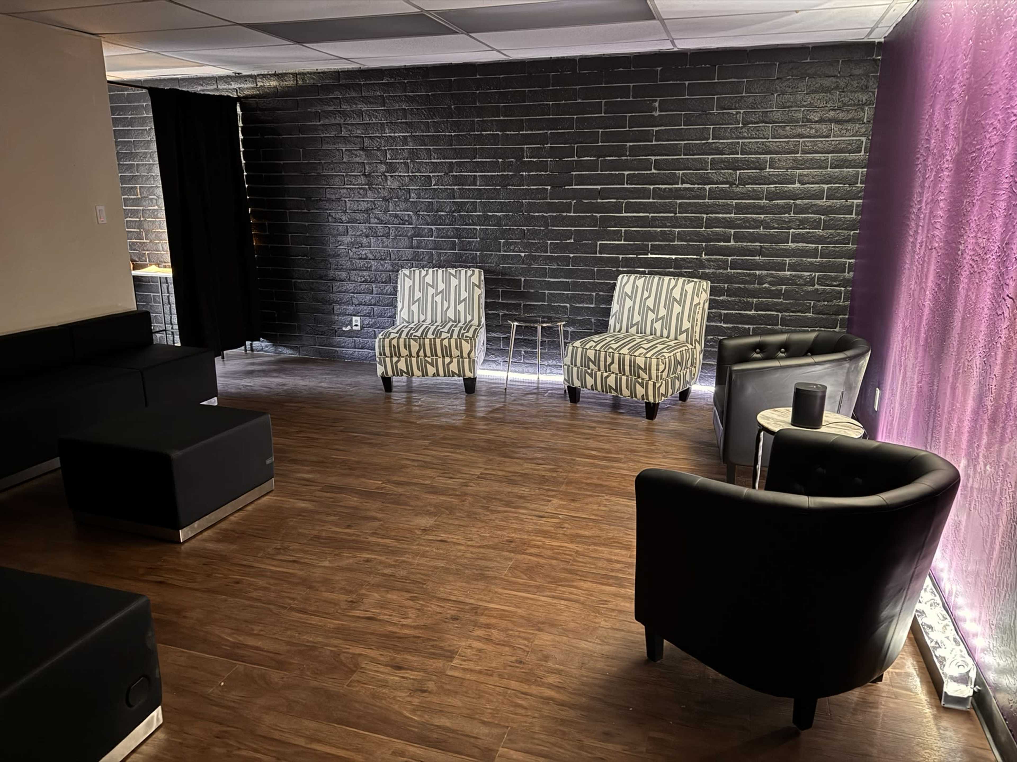 The image shows a modern lounge area with two patterned chairs and two solid black seating options arranged on a wooden floor against a dark brick wall.